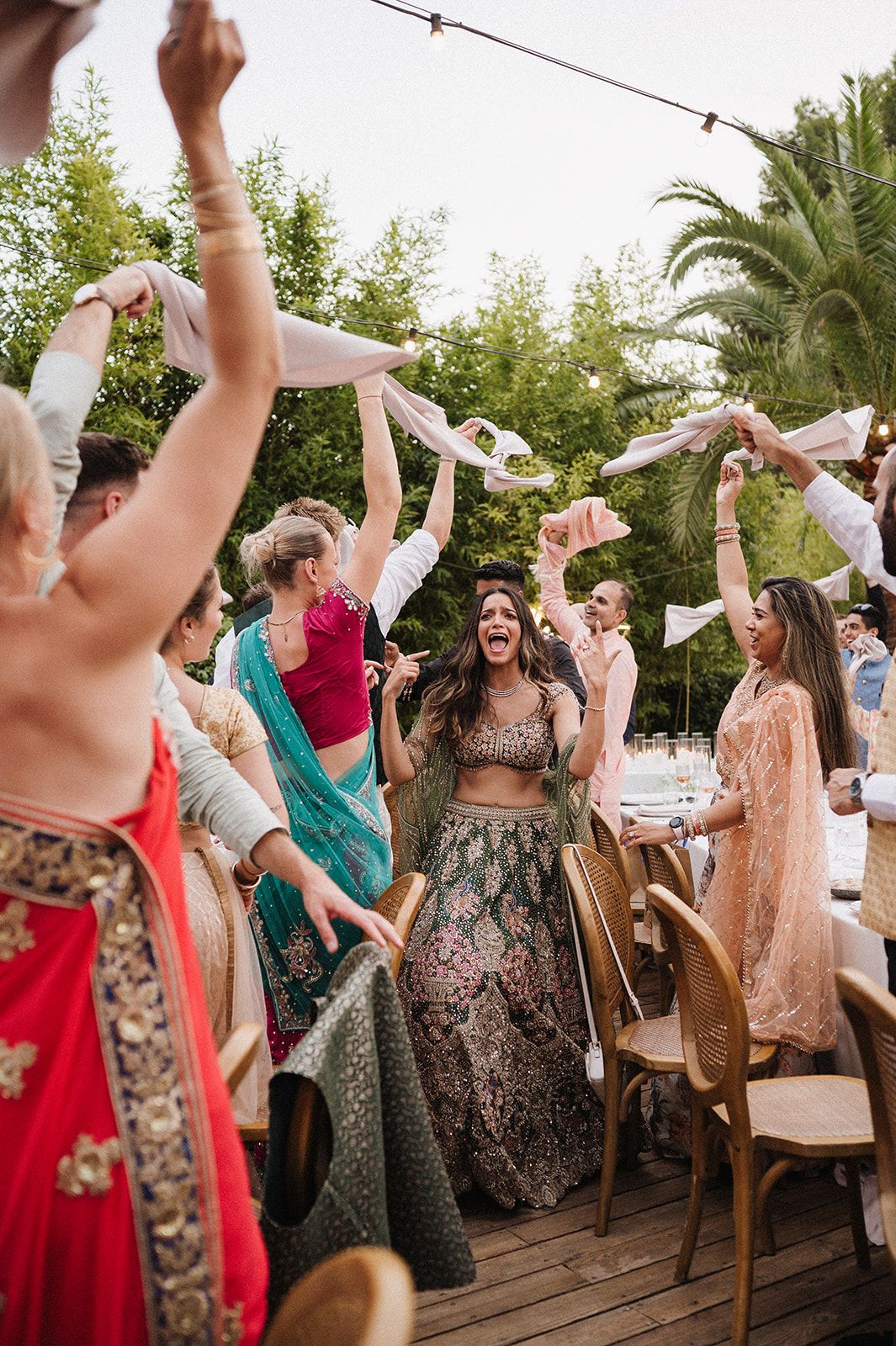 Bride in green lehenga celebrating joyfully as guests wave white scarves at outdoor wedding reception.