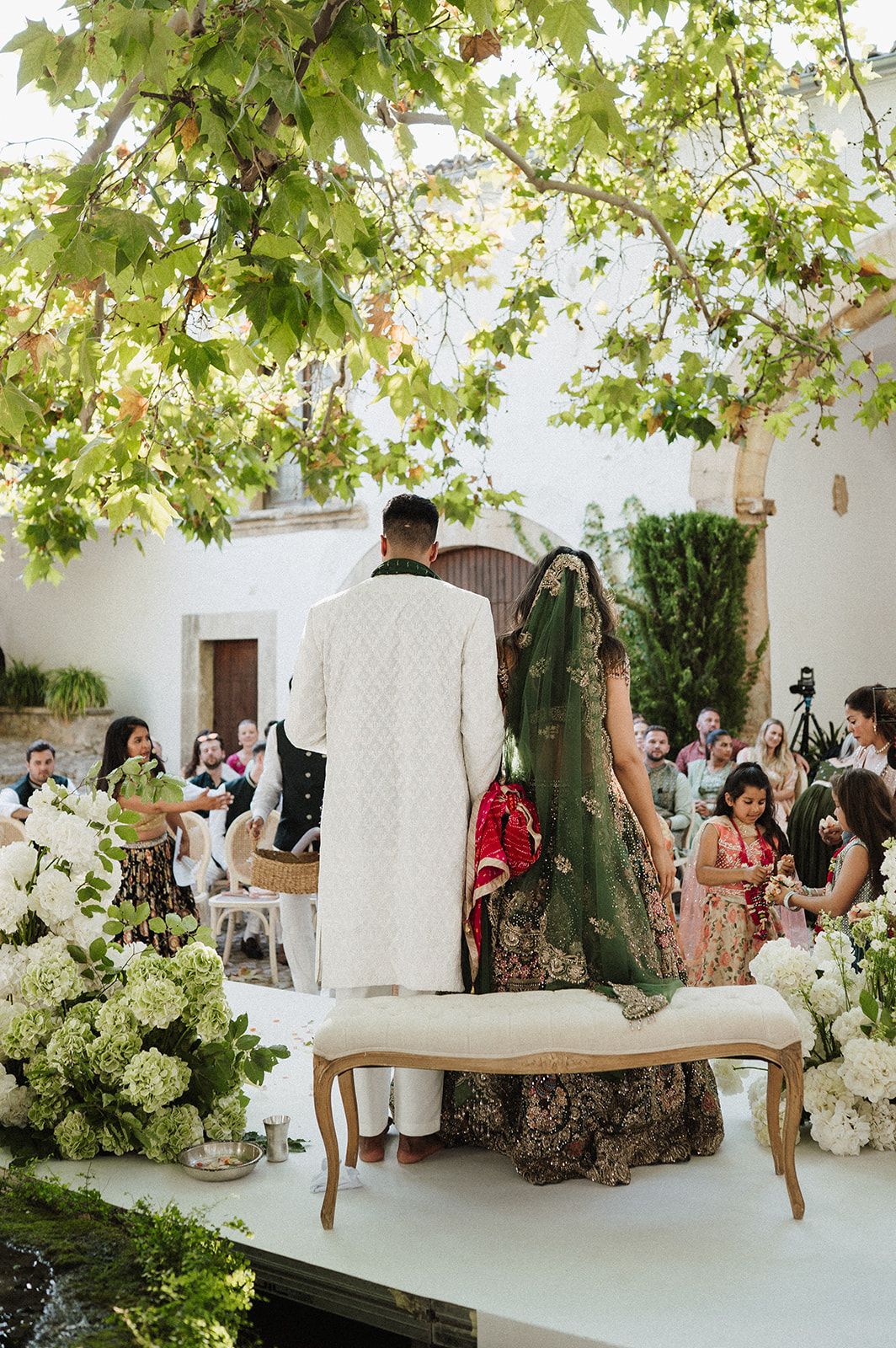 Bride and groom at outdoor Indian wedding ceremony surrounded by white hydrangeas under green trees.