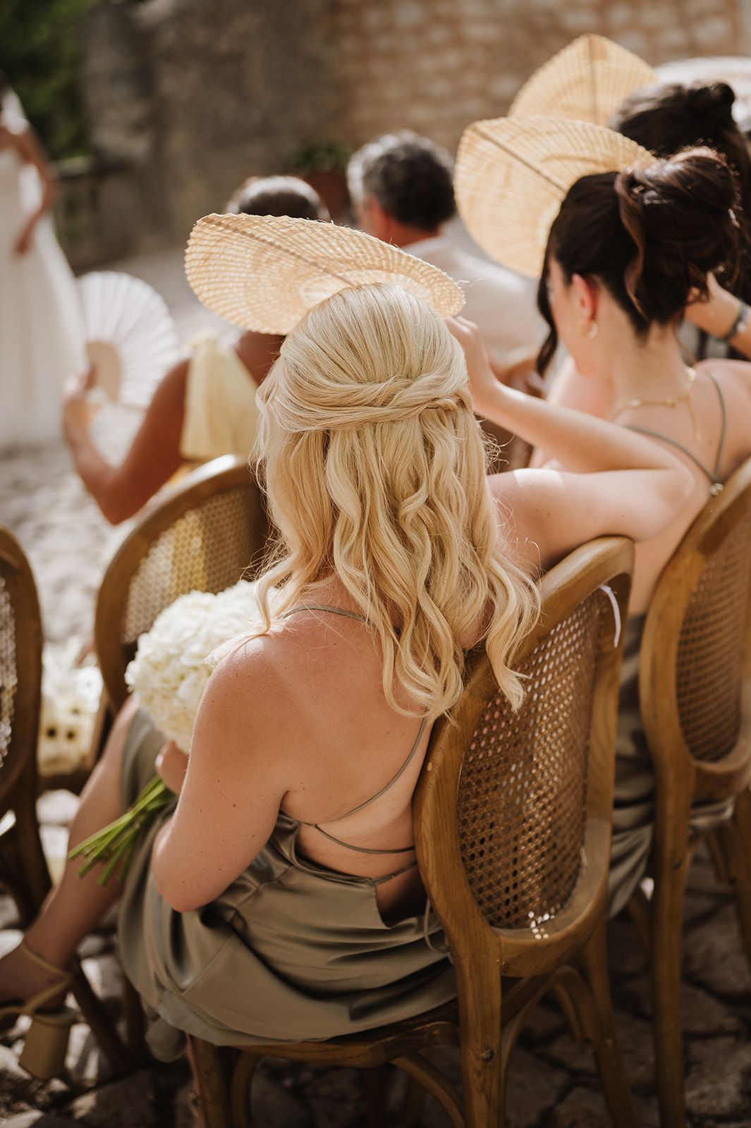 Blonde bridesmaid in sage green dress holds white hydrangeas while seated in rattan chair at outdoor wedding.
