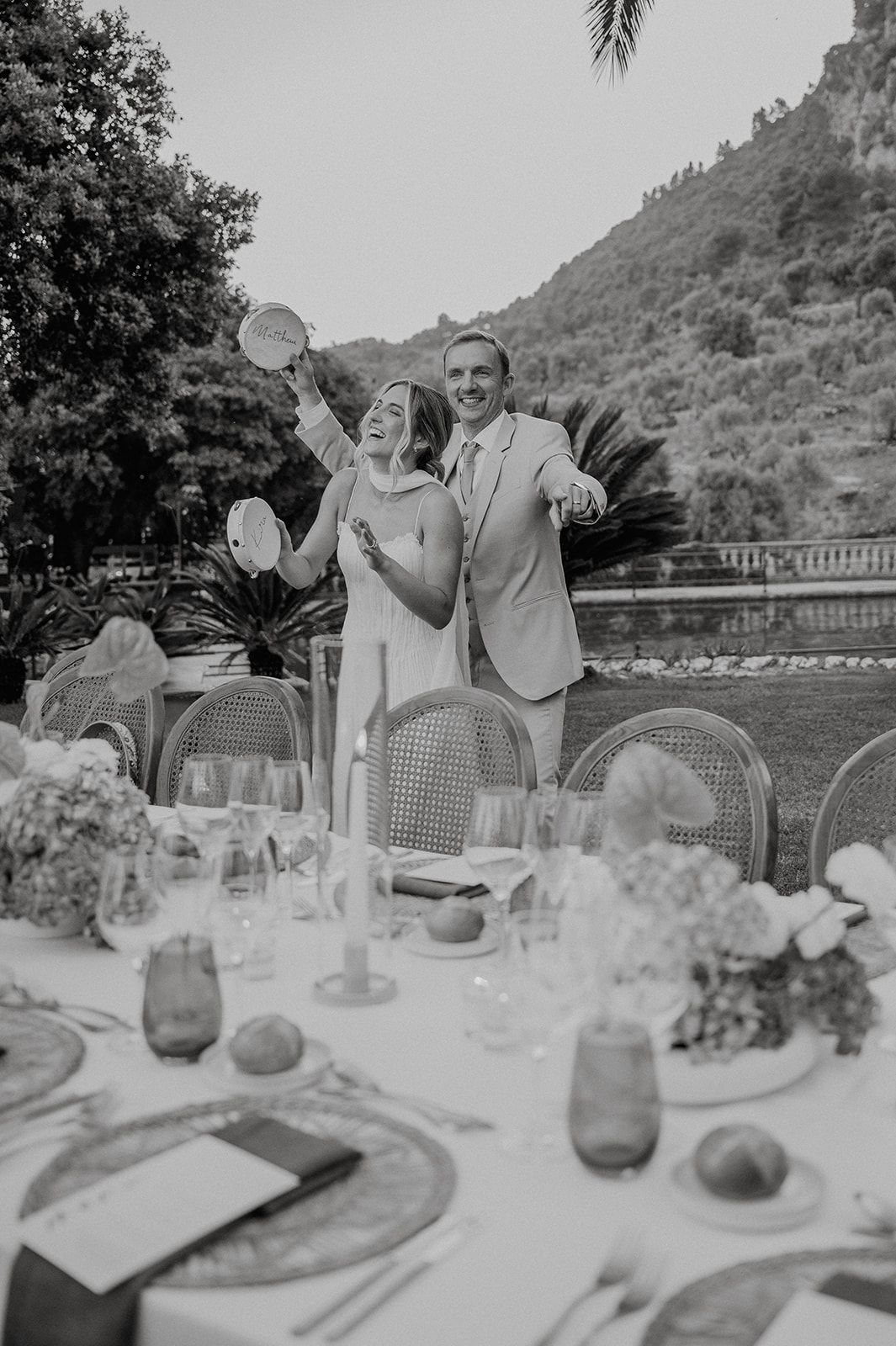 Black and white photo of joyful bride and groom celebrating at elegantly decorated outdoor wedding reception table.