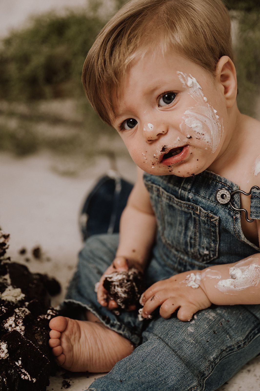 Messy toddler in denim overalls covered in cake and frosting during a first birthday smash cake photoshoot outdoors.