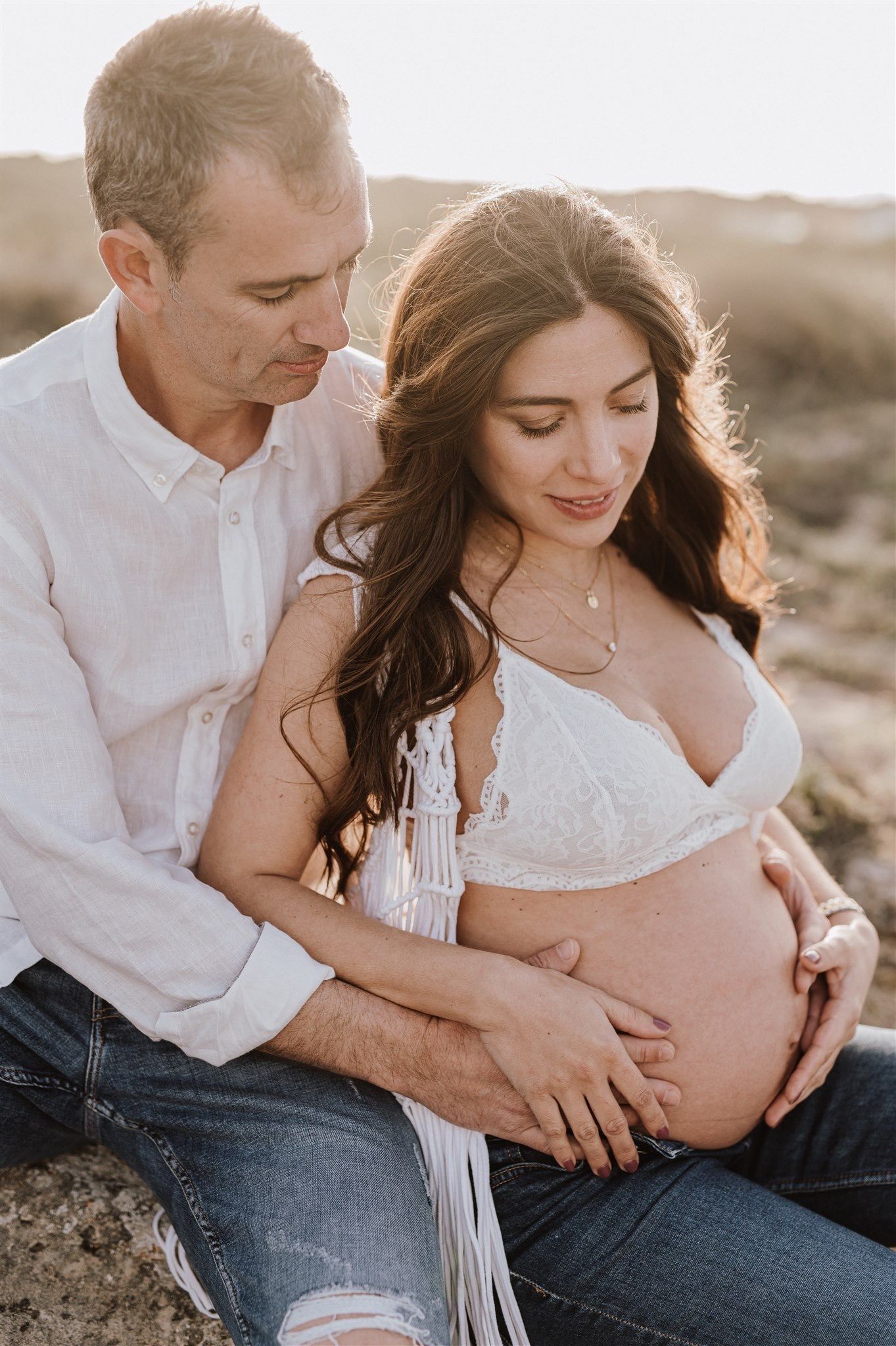Couple sharing a tender maternity moment outdoors, man embracing pregnant woman in white lace top and jeans at golden hour.