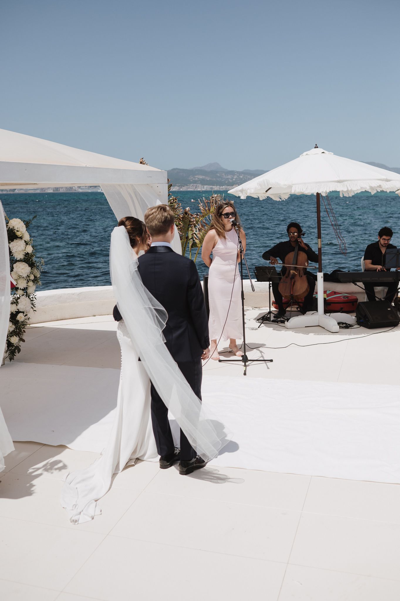 Outdoor seaside wedding ceremony with bride and groom at altar, live cello music and singer performing on white terrace.