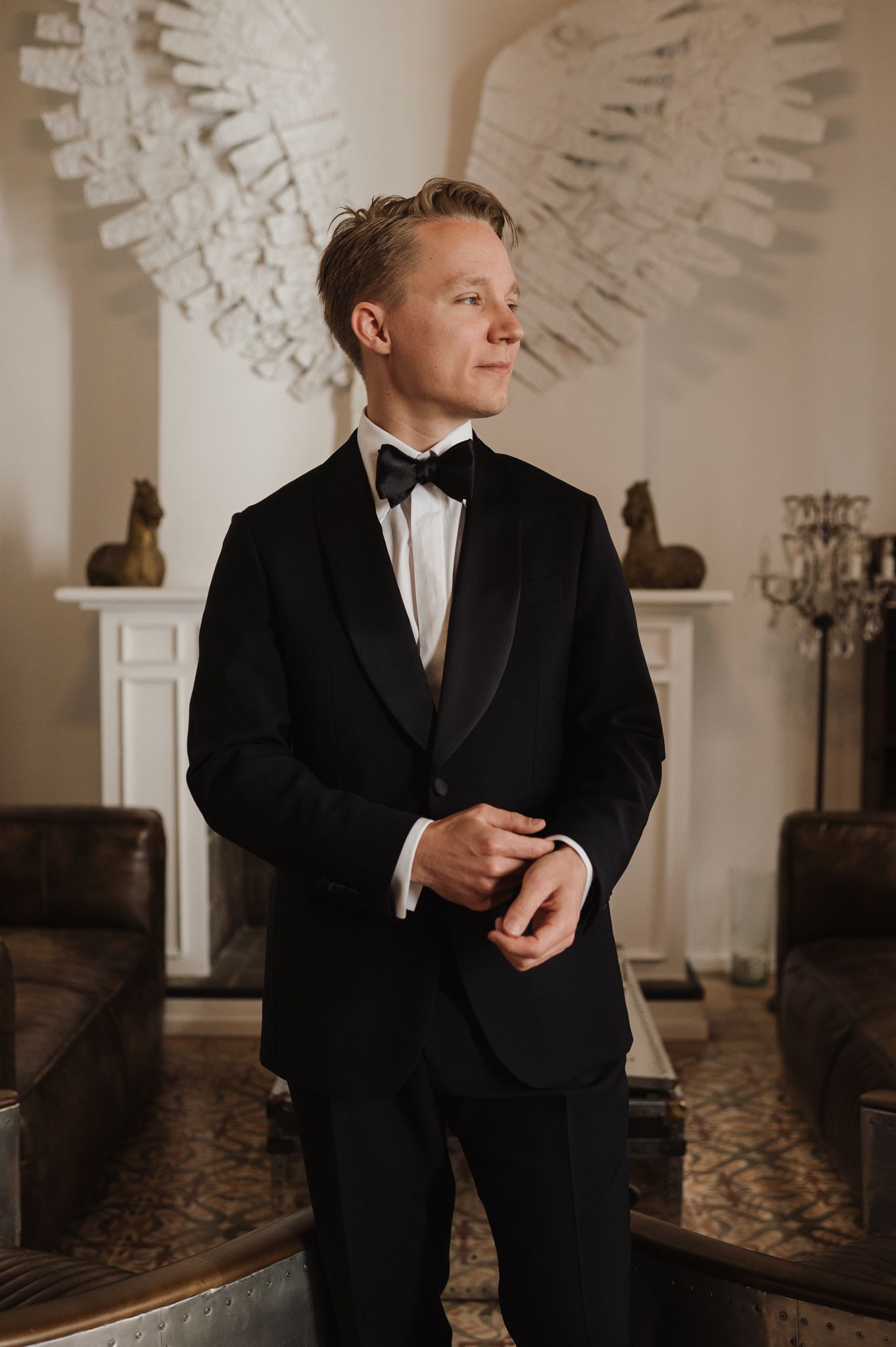 Handsome groom in black tuxedo and bow tie adjusting his jacket in a luxurious hotel room.