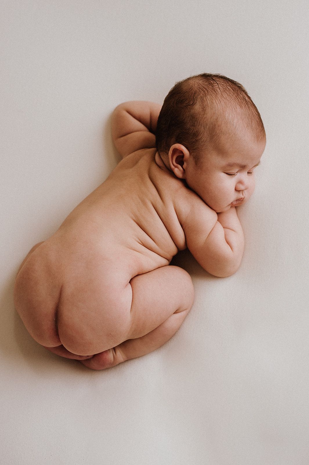 Peaceful newborn baby sleeping curled up in fetal position on soft white background, showing chubby skin folds.
