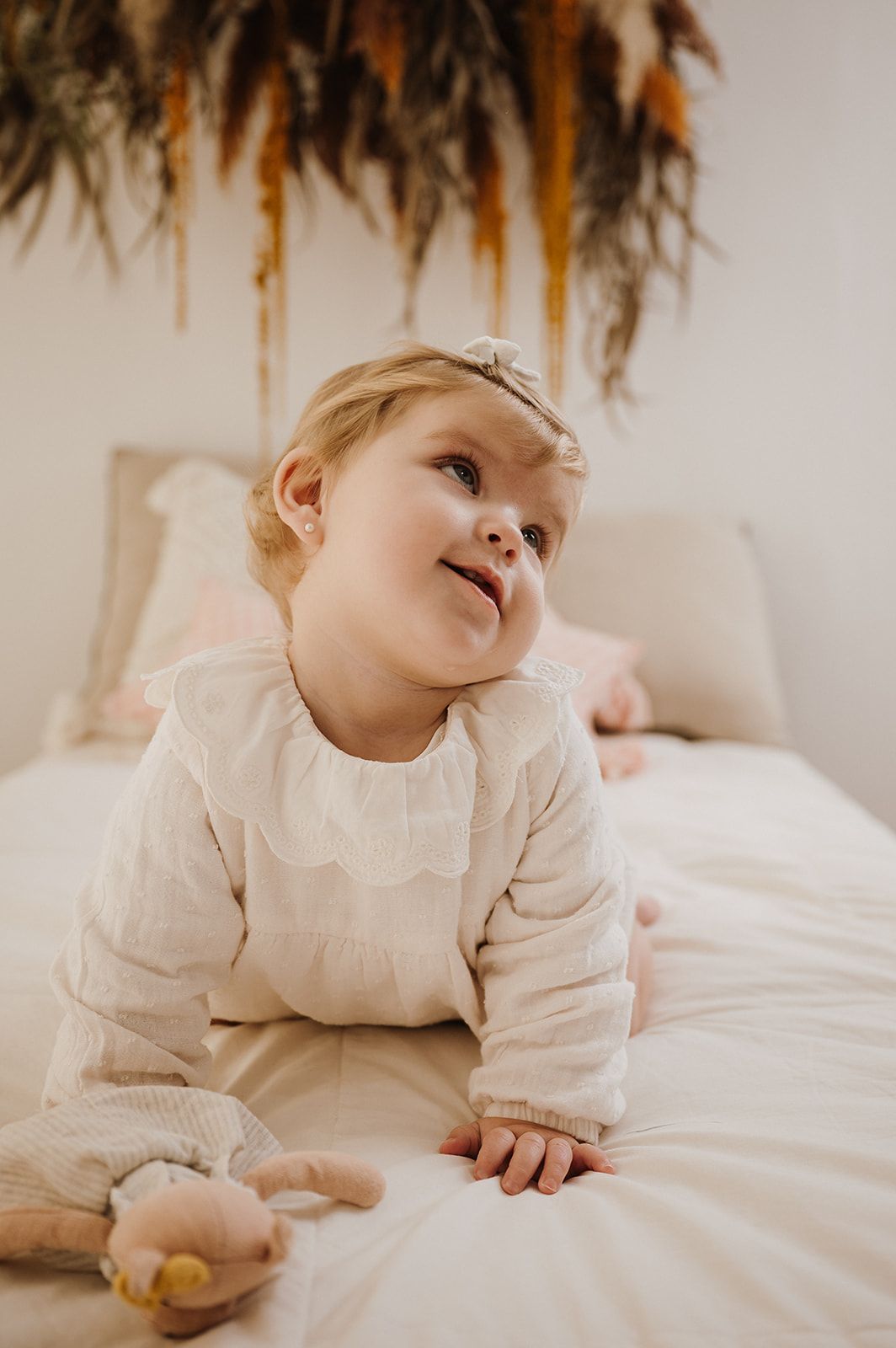 Toddler girl in cream dress with headband lying on white bed smiling.