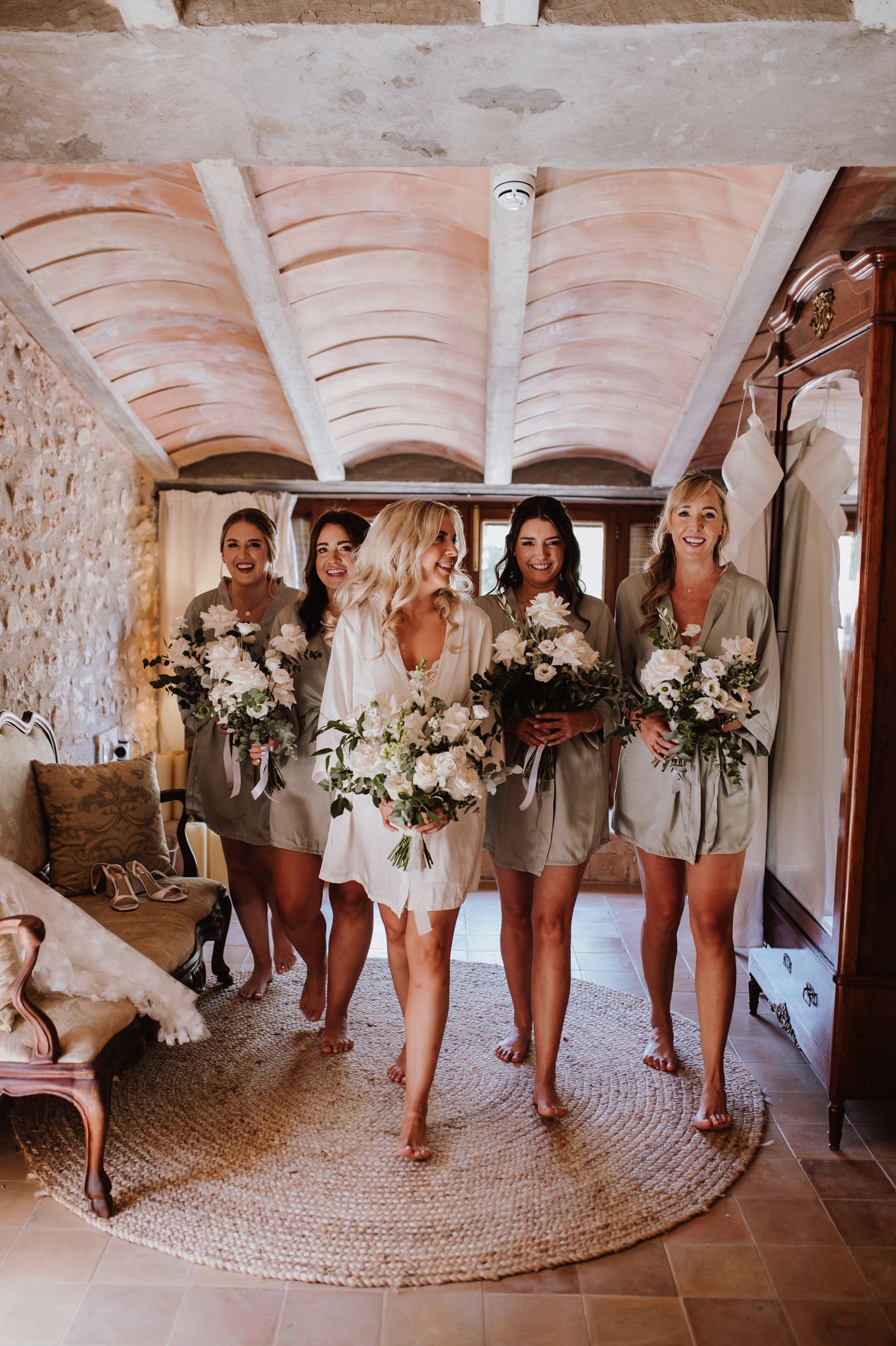 Bride in white robe with bridesmaids in sage rompers holding white floral bouquets in a rustic stone venue.