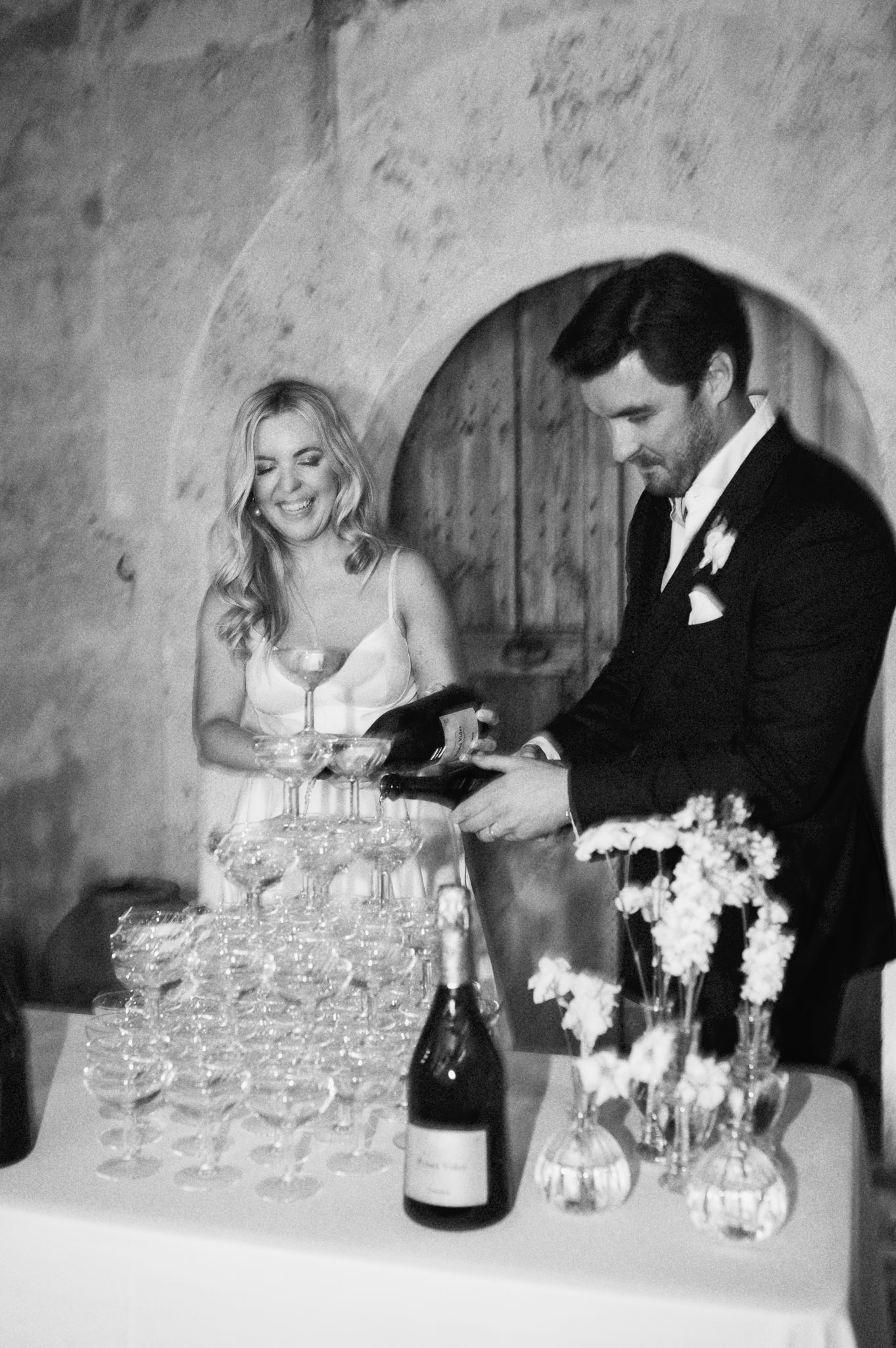 Black and white photo of bride and groom pouring champagne tower at elegant wedding reception.