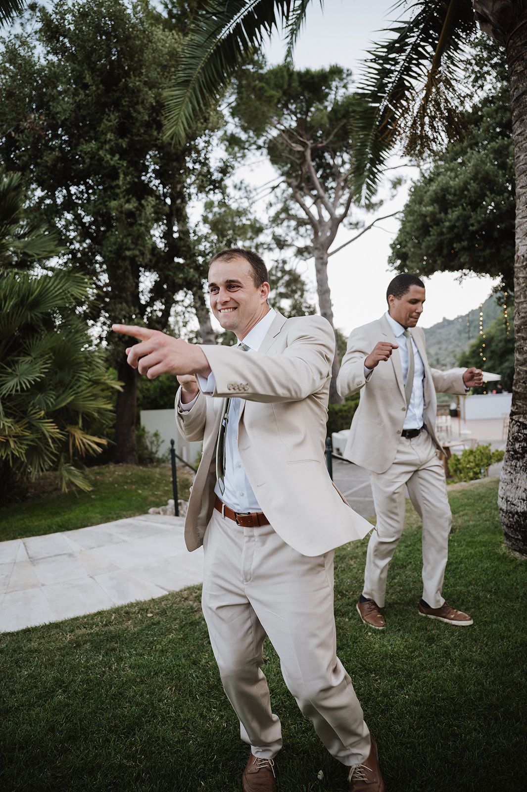 Two groomsmen in tan suits dancing and pointing outdoors near palm trees during a wedding celebration.
