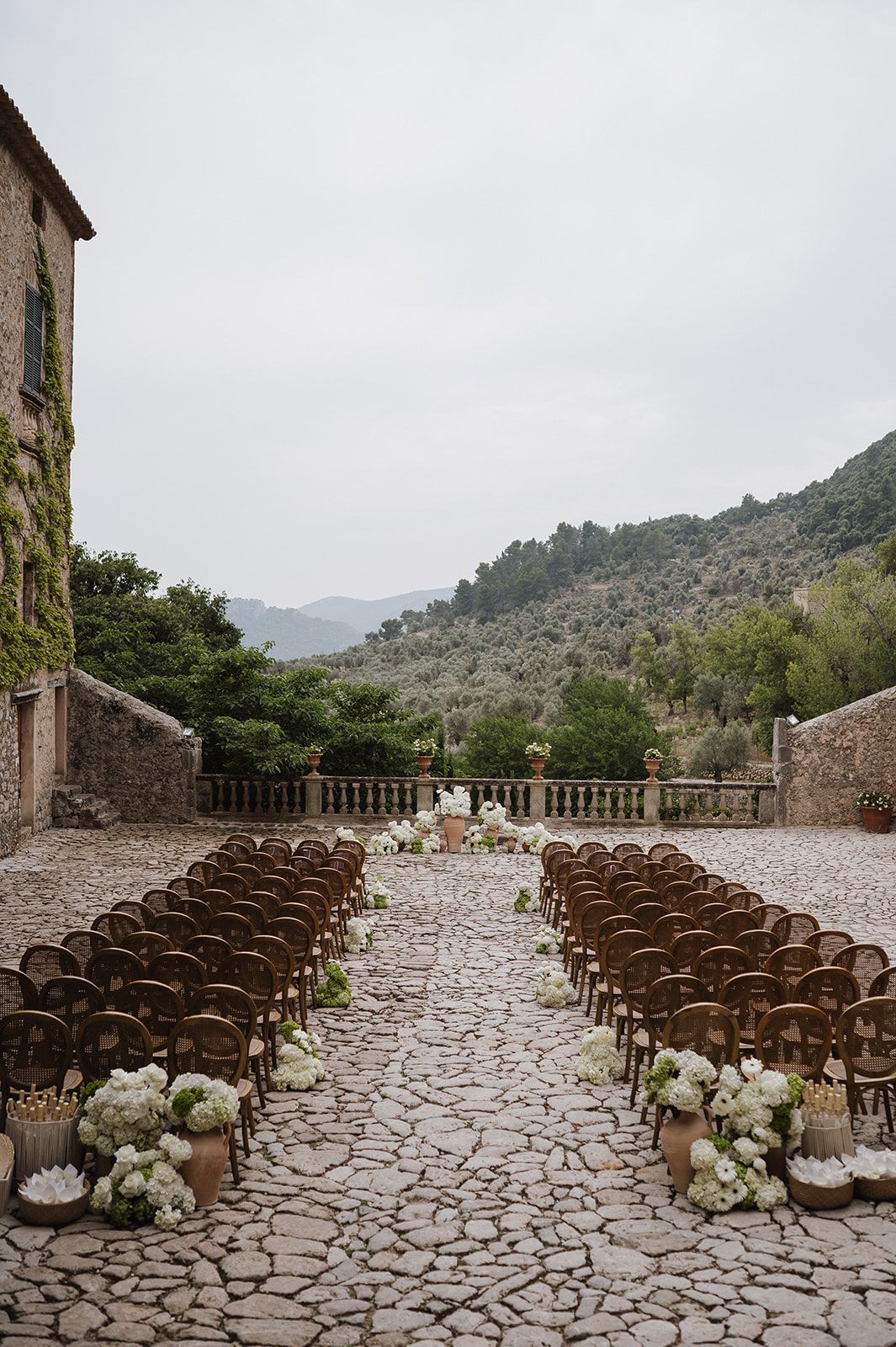 Elegant outdoor wedding ceremony setup with wooden chairs, white floral arrangements, and mountain backdrop.