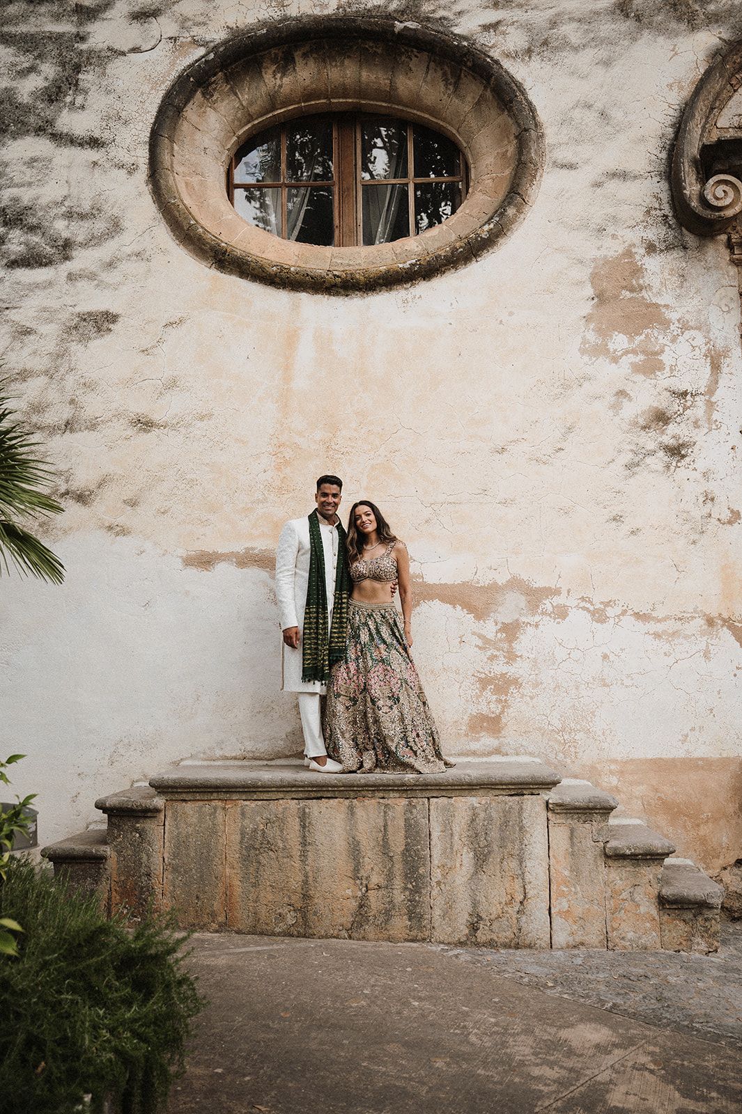 Indian wedding couple posing on stone steps before a rustic wall with circular window.
