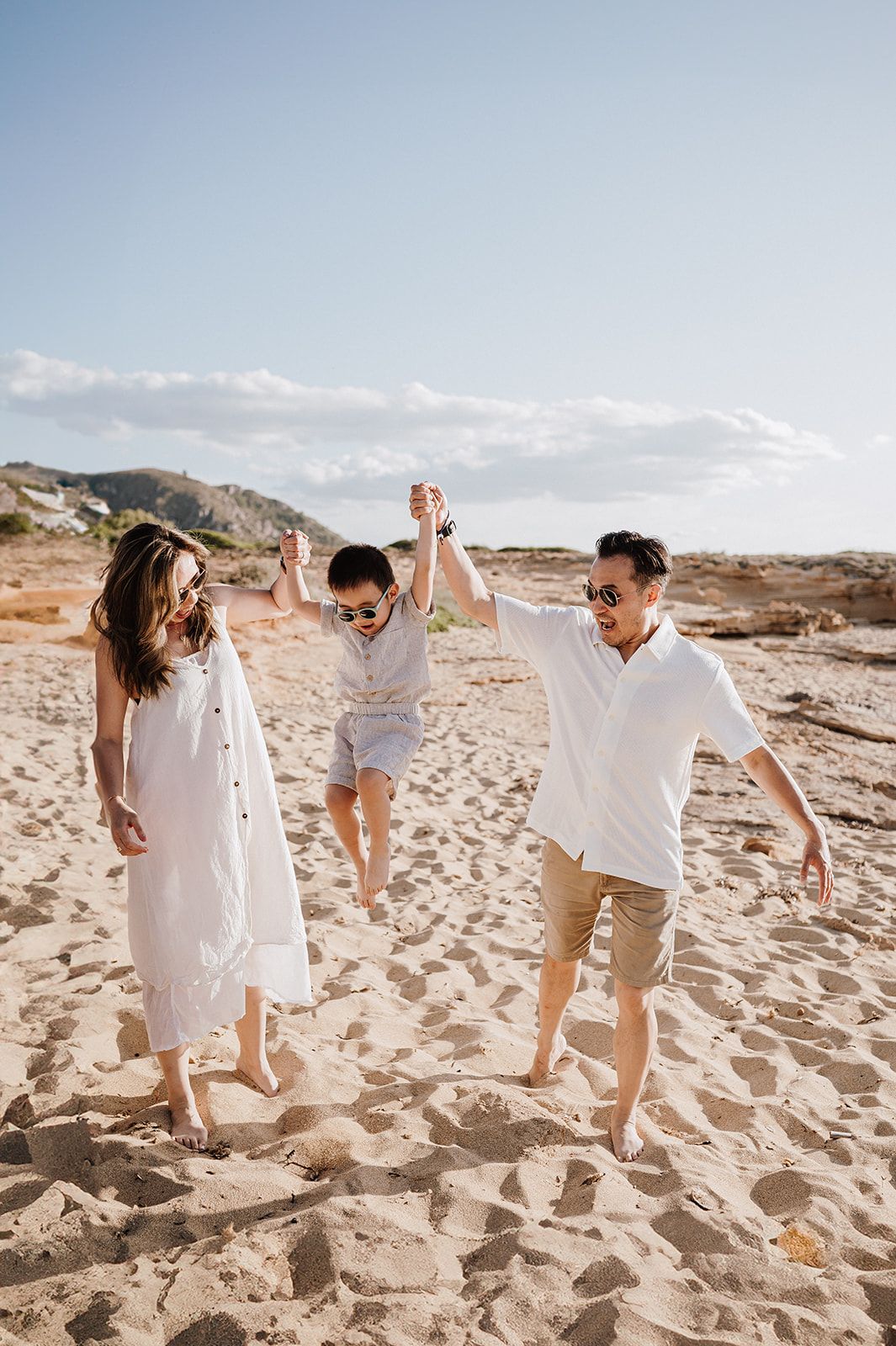 Family of three in white outfits joyfully swinging child on sandy beach under clear blue sky.