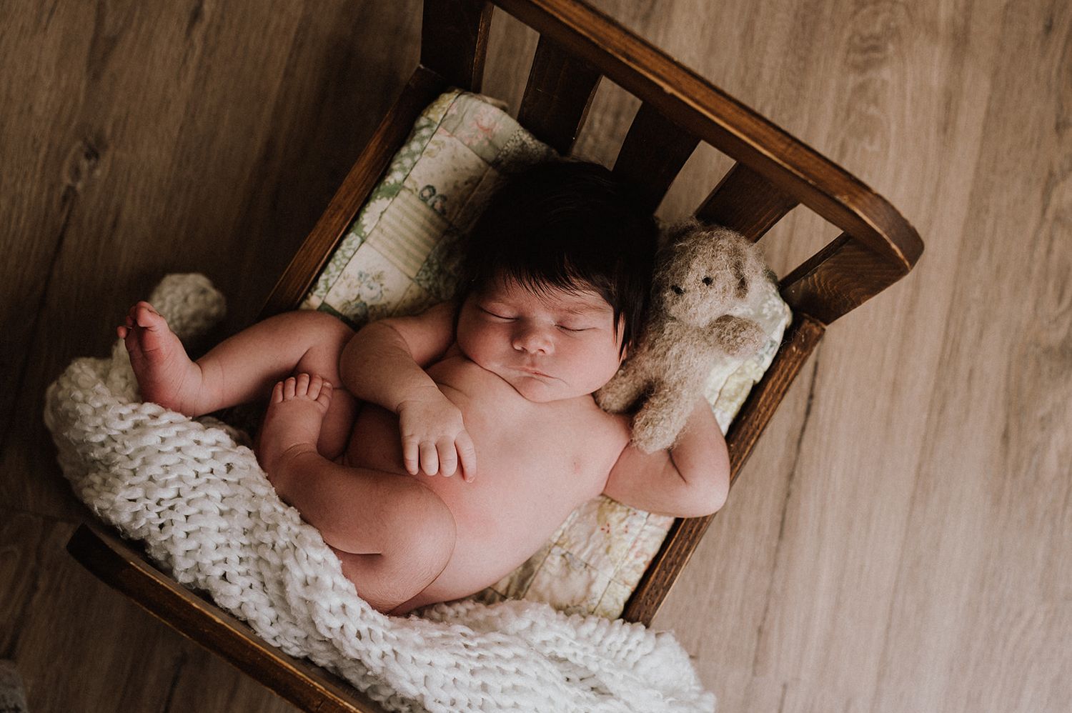 Newborn baby sleeping peacefully in a tiny wooden bed with a knit blanket and stuffed teddy bear.