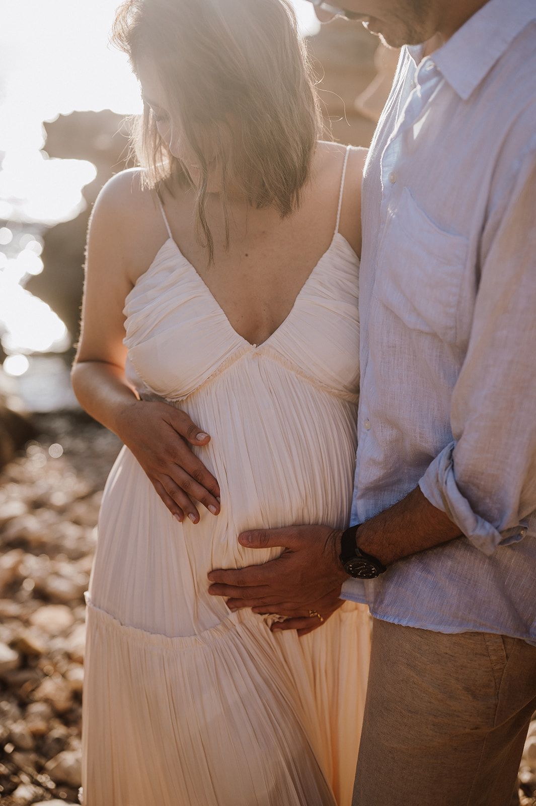 Pregnant woman in white dress with partner tenderly cradling baby bump on rocky beach at golden hour.