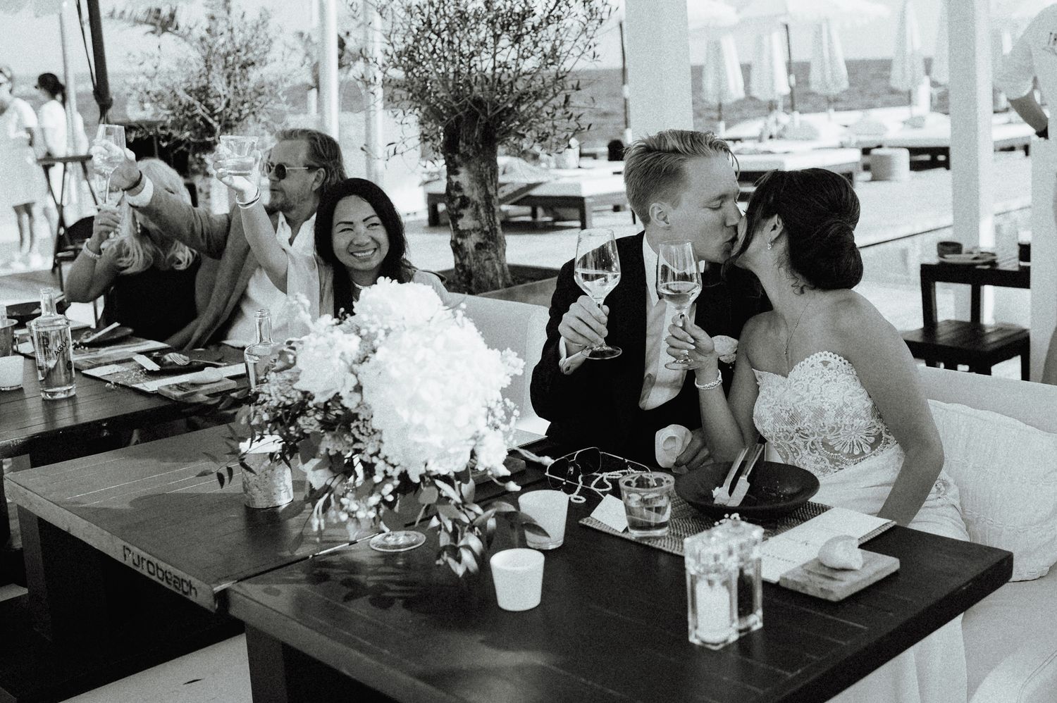 Black and white photo of bride and groom sharing a kiss at wedding reception table with flowers.