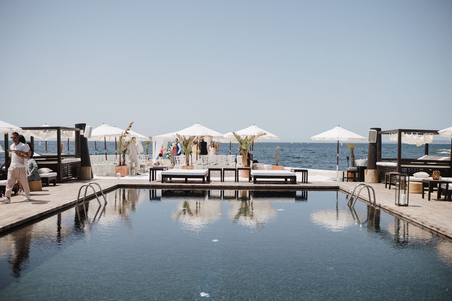 Elegant seaside pool venue with white umbrellas and ocean views set up for a wedding ceremony.