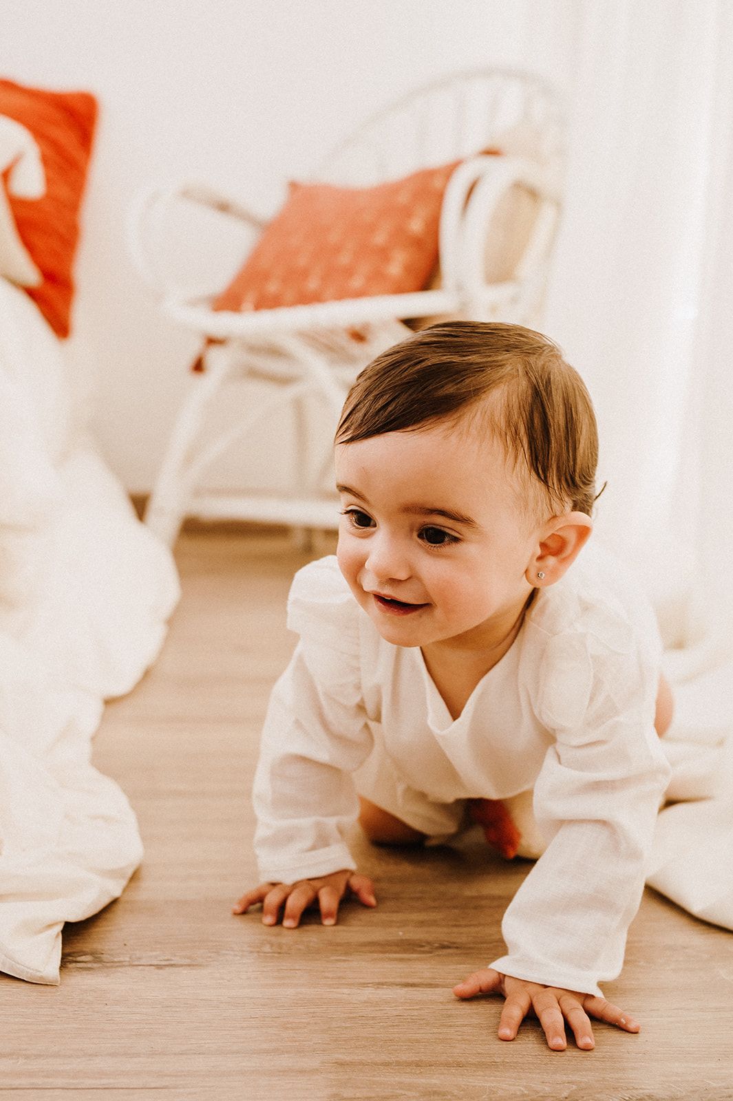 A happy baby in a white outfit crawls on a hardwood floor, smiling, with a white rattan chair in the background.