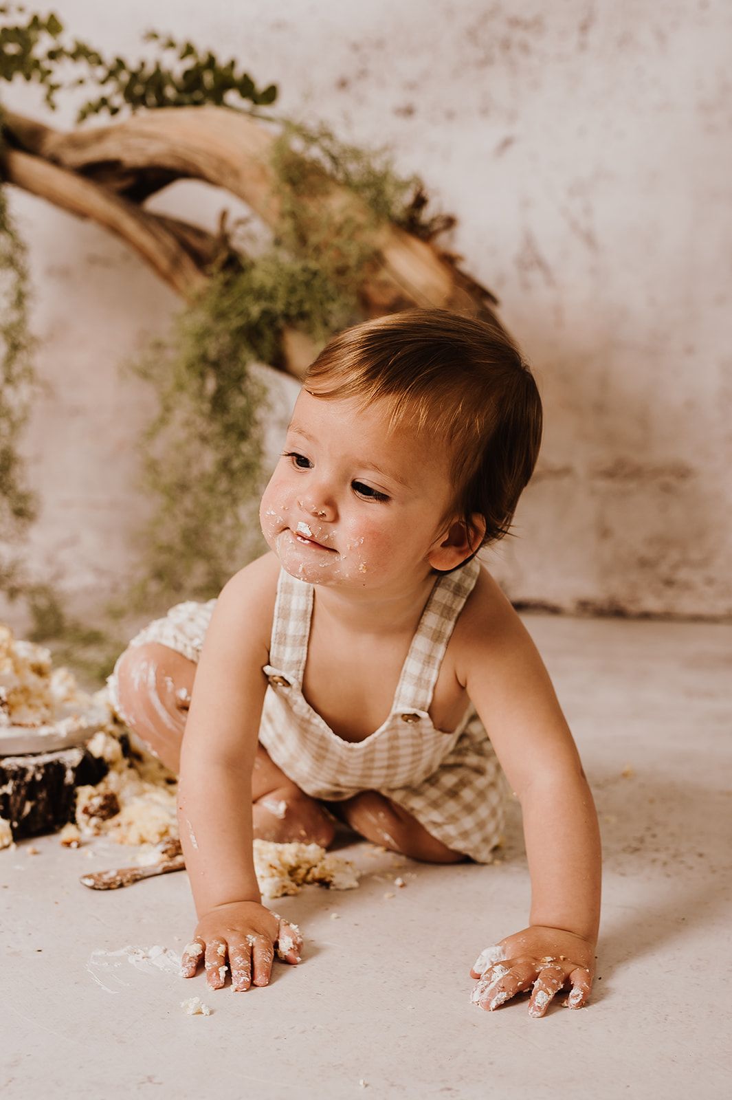 A baby in gingham overalls crawls on the floor with cake smash mess on hands and face during a first birthday photoshoot.