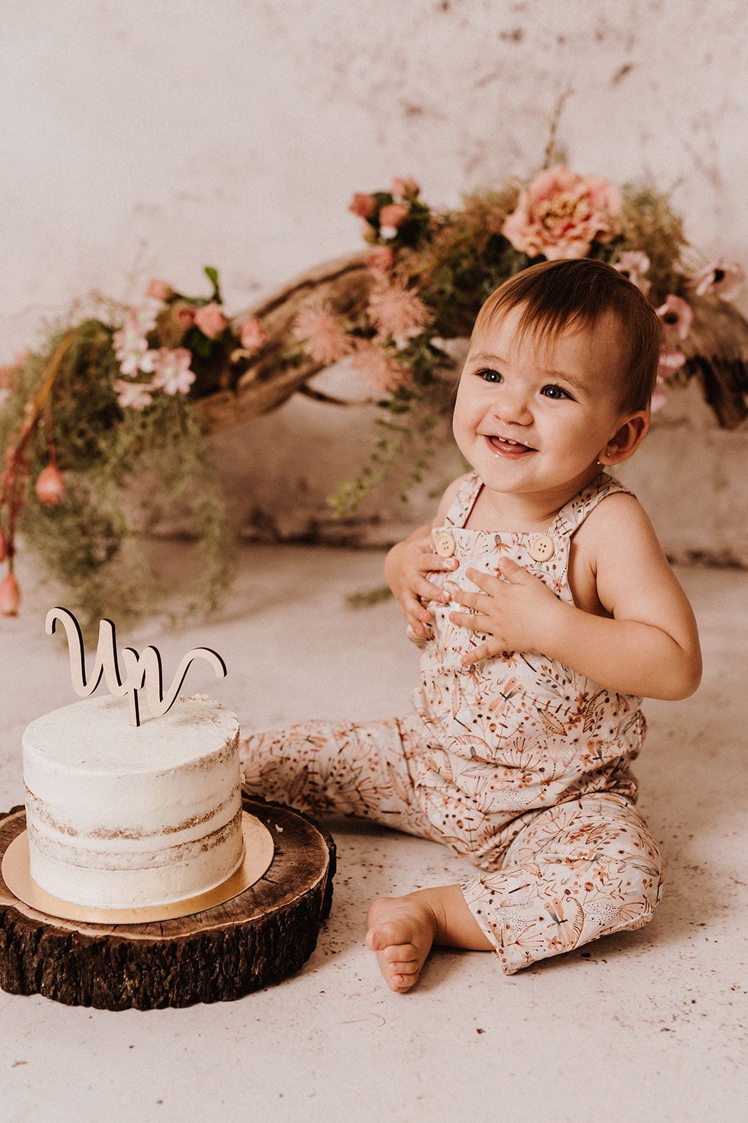 Smiling baby girl in floral romper sitting next to a white cake topper at a first birthday cake smash photo session.