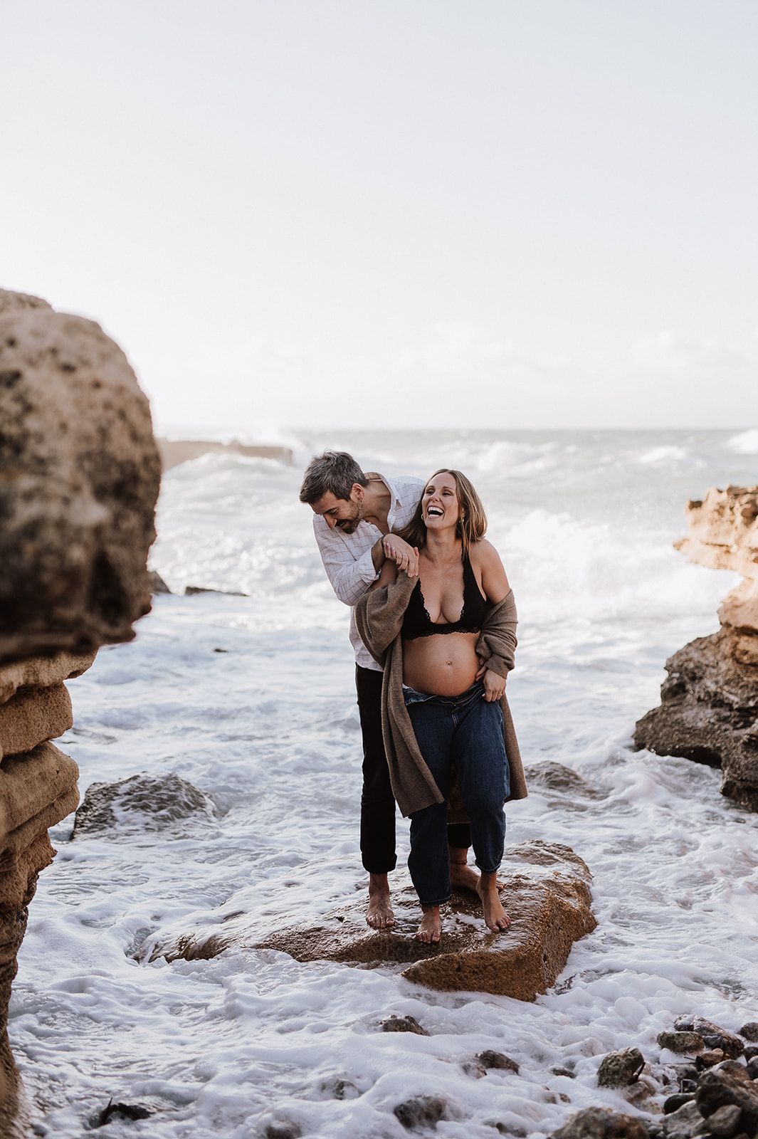 Pregnant couple laughing together on rocky ocean shore, waves splashing around their feet during a maternity photo session.