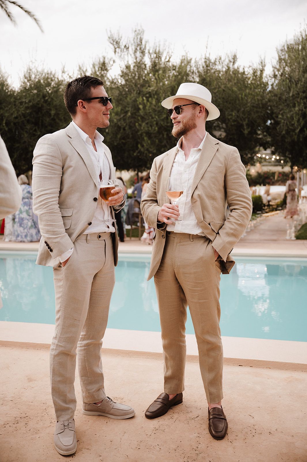 Two men in beige linen suits holding drinks by a pool at an outdoor summer wedding reception.