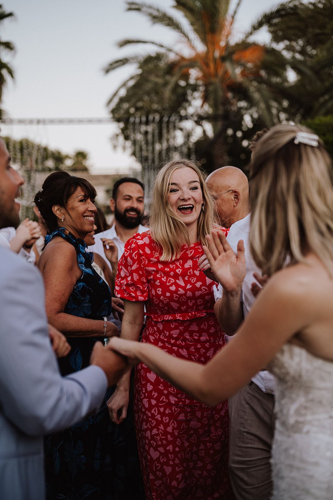 Laughing wedding guest in red floral dress celebrates with bride and other guests at outdoor reception.