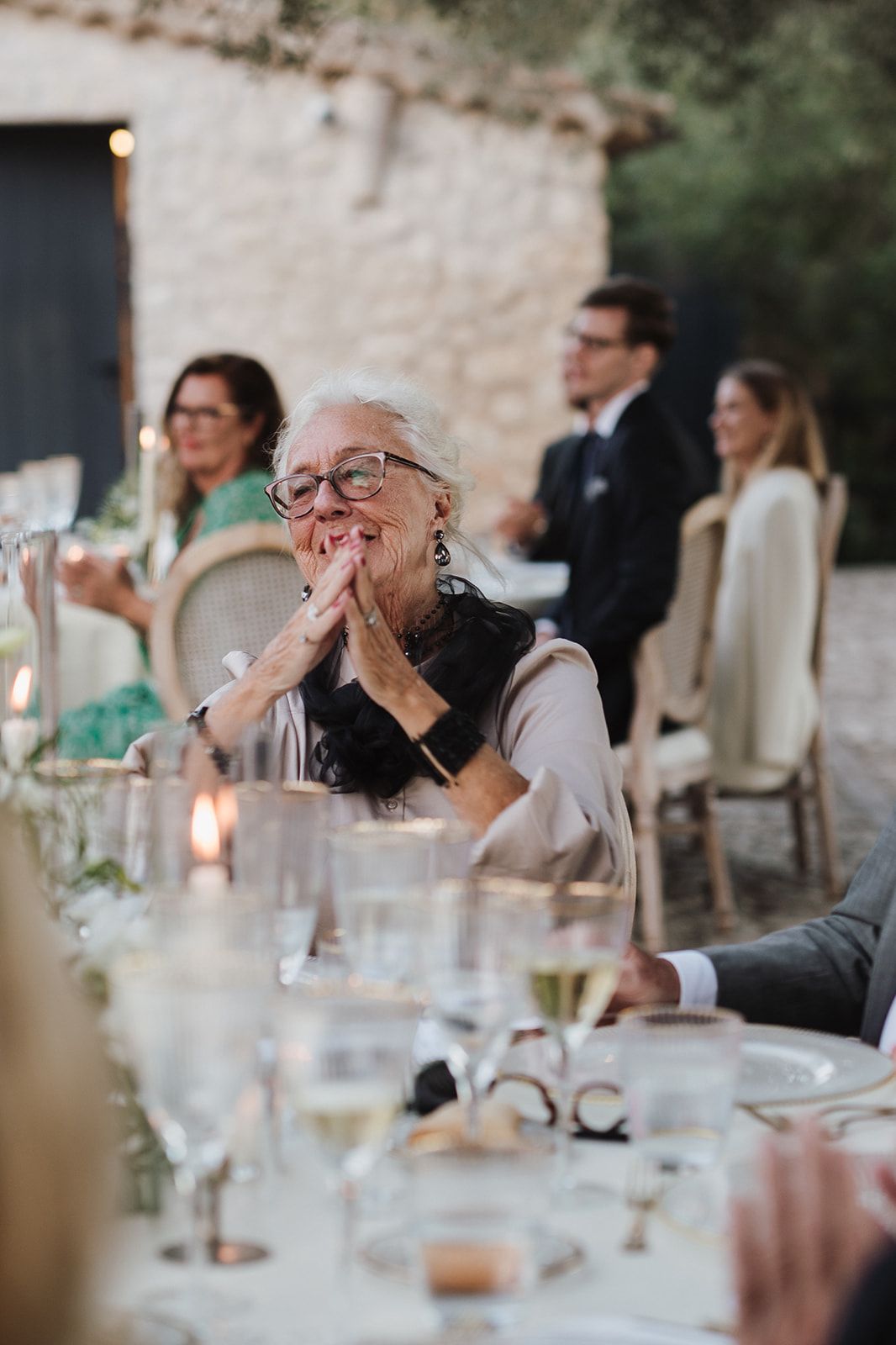 Elderly wedding guest smiling with hands clasped at elegant outdoor reception dinner.