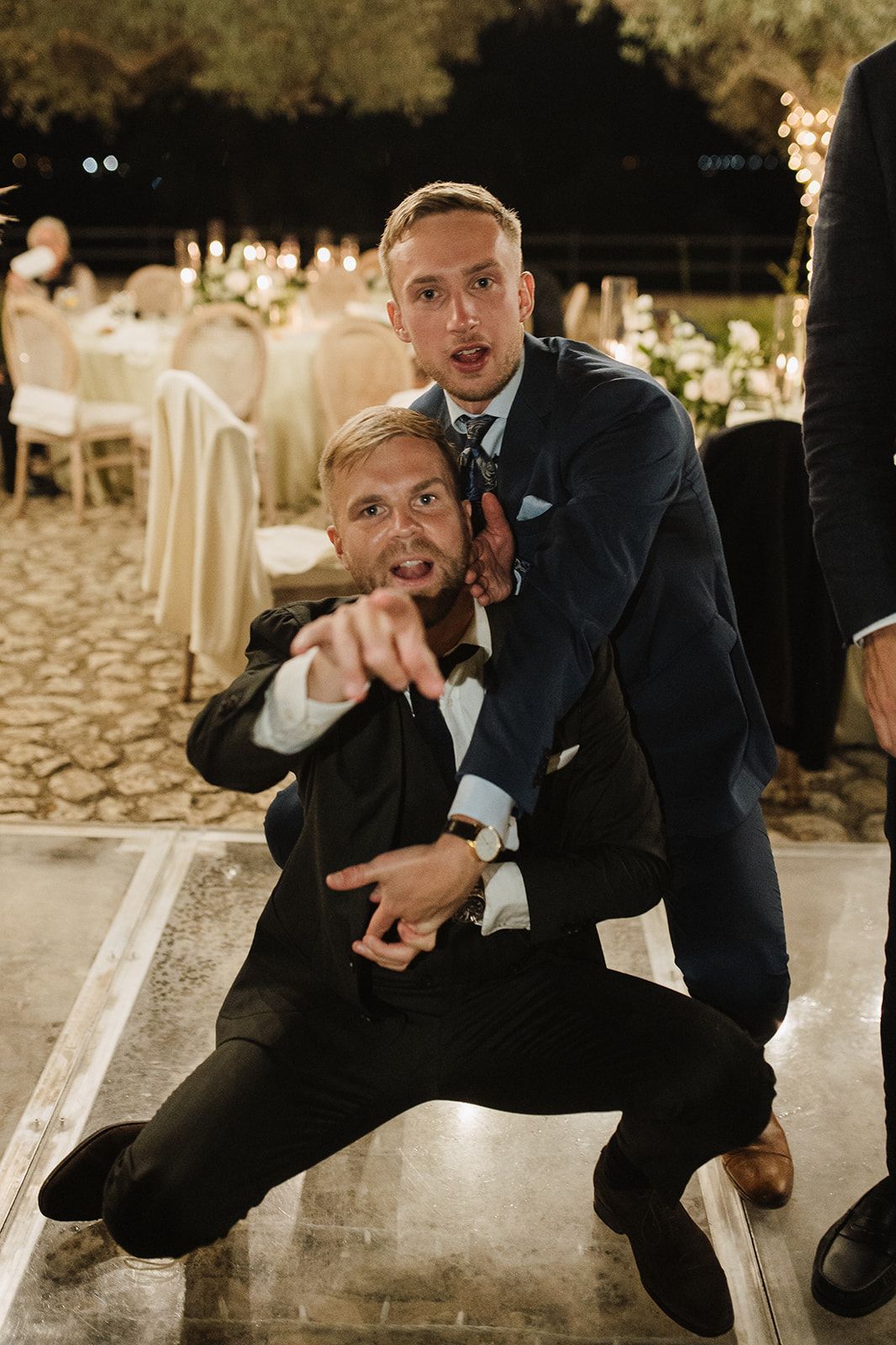 Two male wedding guests pose playfully on the dance floor during evening reception.