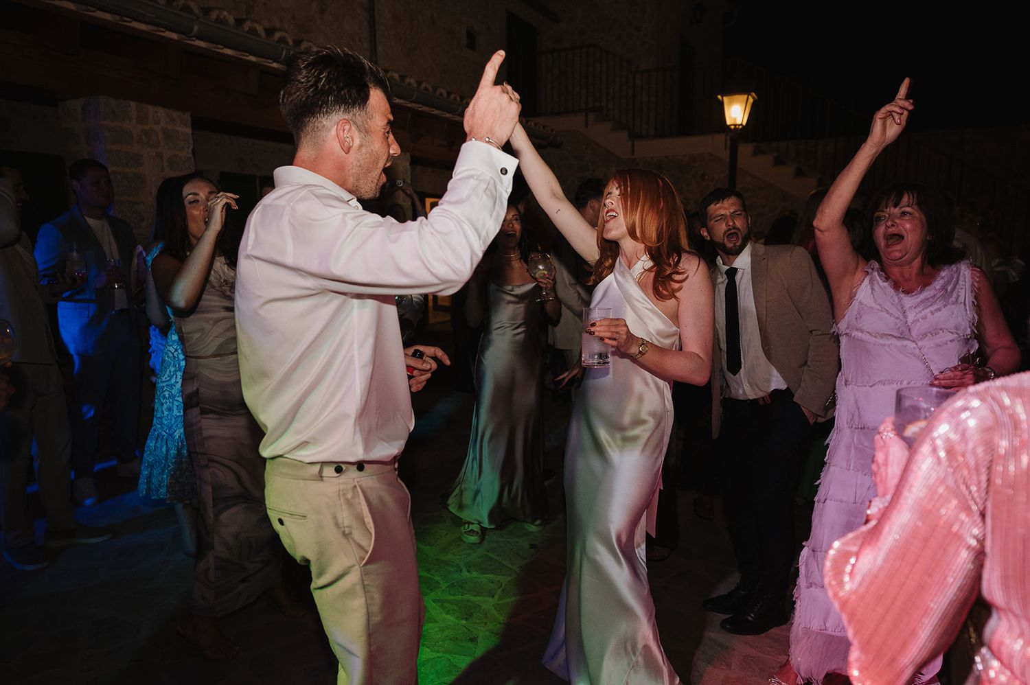 Bride in white satin dress dancing energetically with groom and guests at outdoor wedding reception.