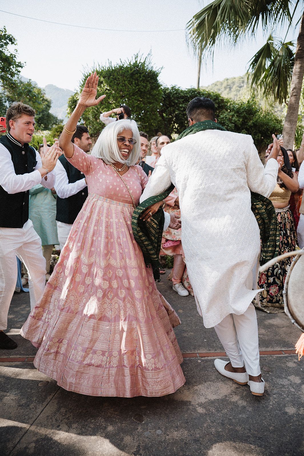 Elderly woman in pink lehenga dancing joyfully at an Indian wedding celebration outdoors.