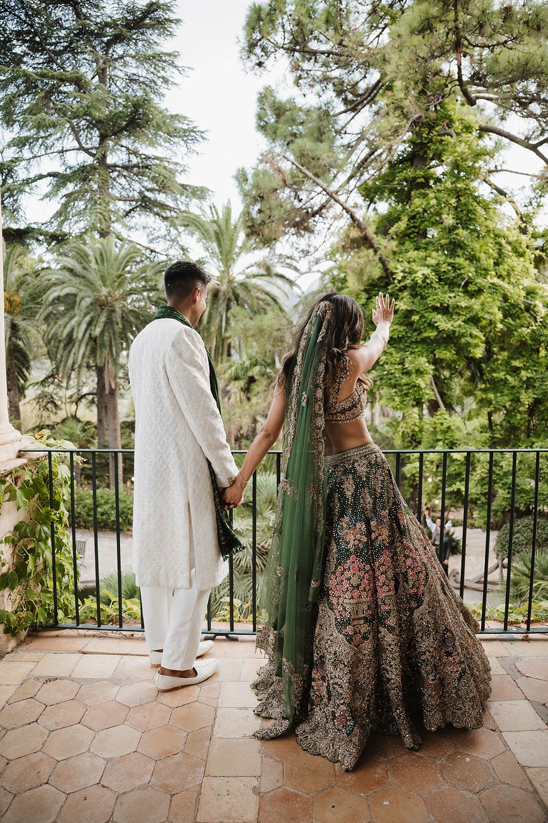 Bride in green embroidered lehenga and groom in white sherwani holding hands on a balcony.
