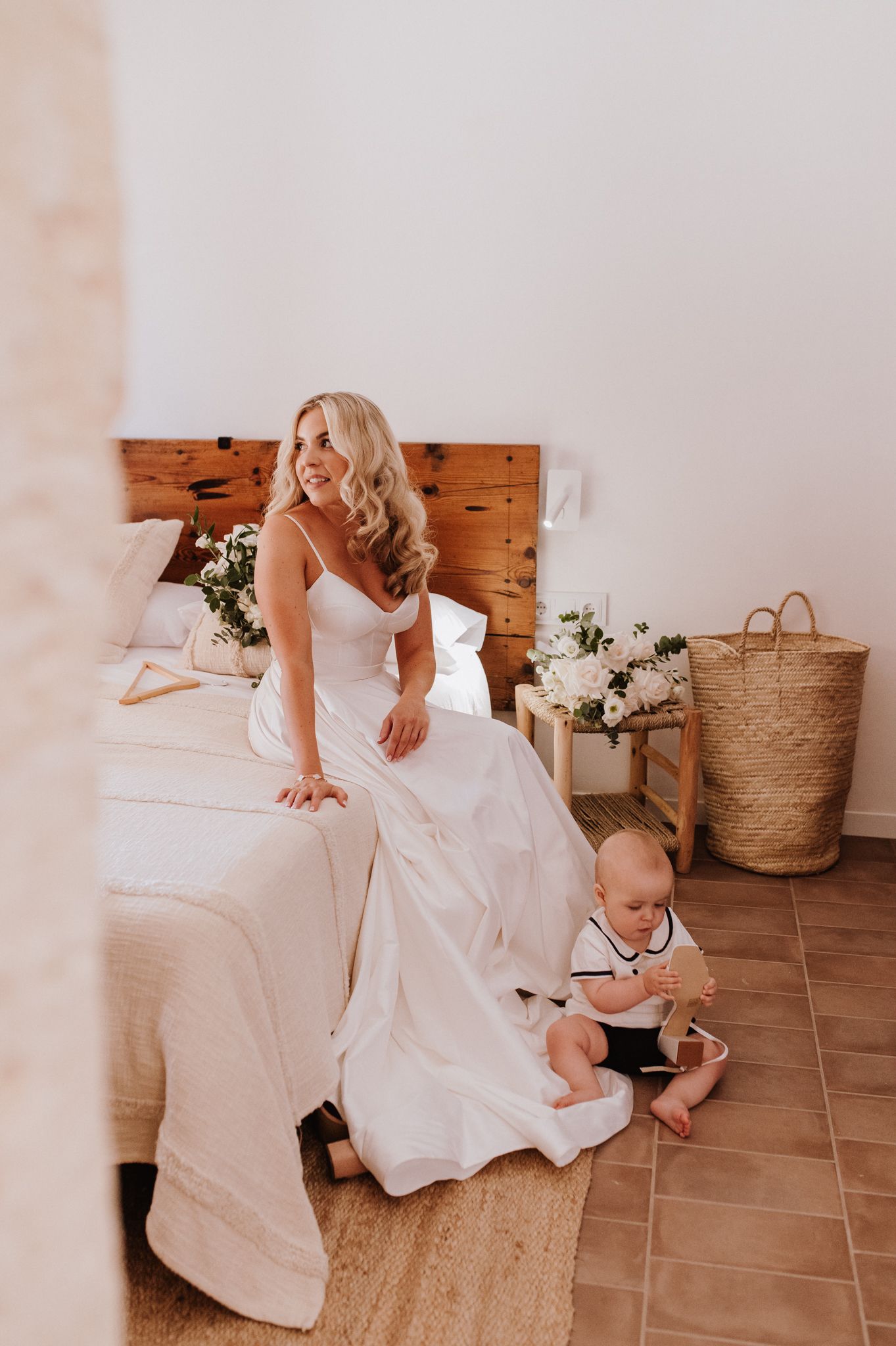 Bride in white wedding dress sitting on bed beside a baby in formal attire in a bright rustic bedroom.