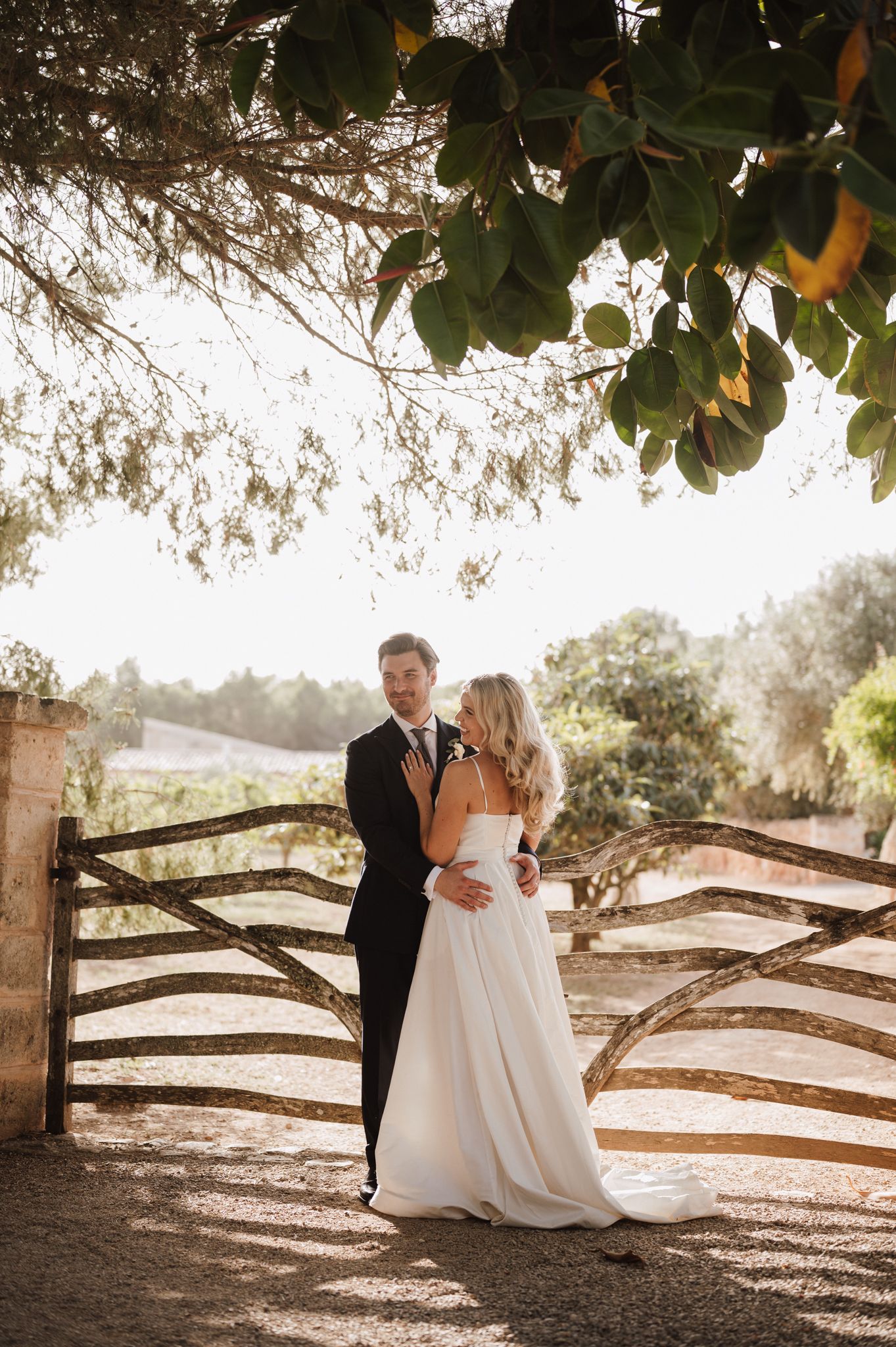 Bride and groom sharing romantic moment by wooden fence in golden sunset light at outdoor wedding.