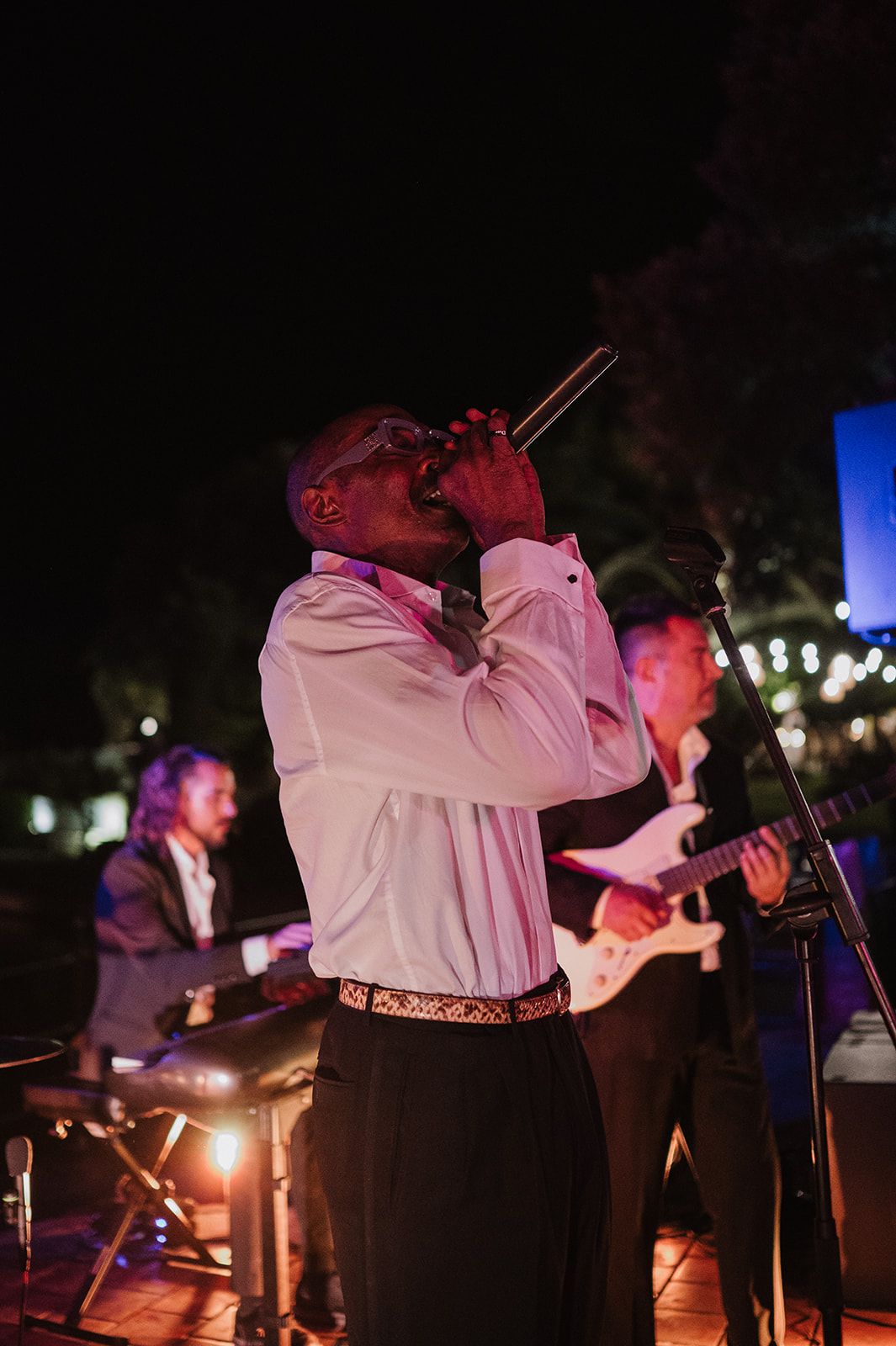 Live band performer singing into microphone at outdoor wedding reception under evening lights.