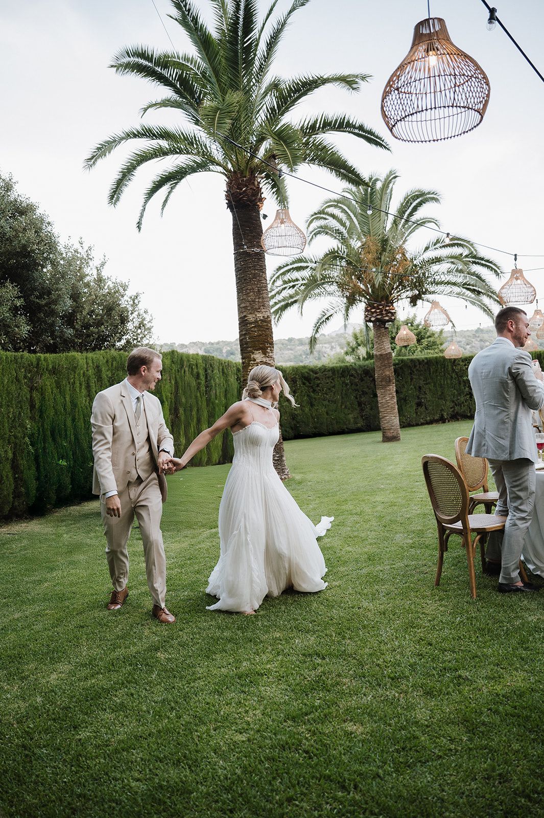 Bride in white flowing gown and groom in beige suit hold hands dancing on green lawn under palm trees at reception.