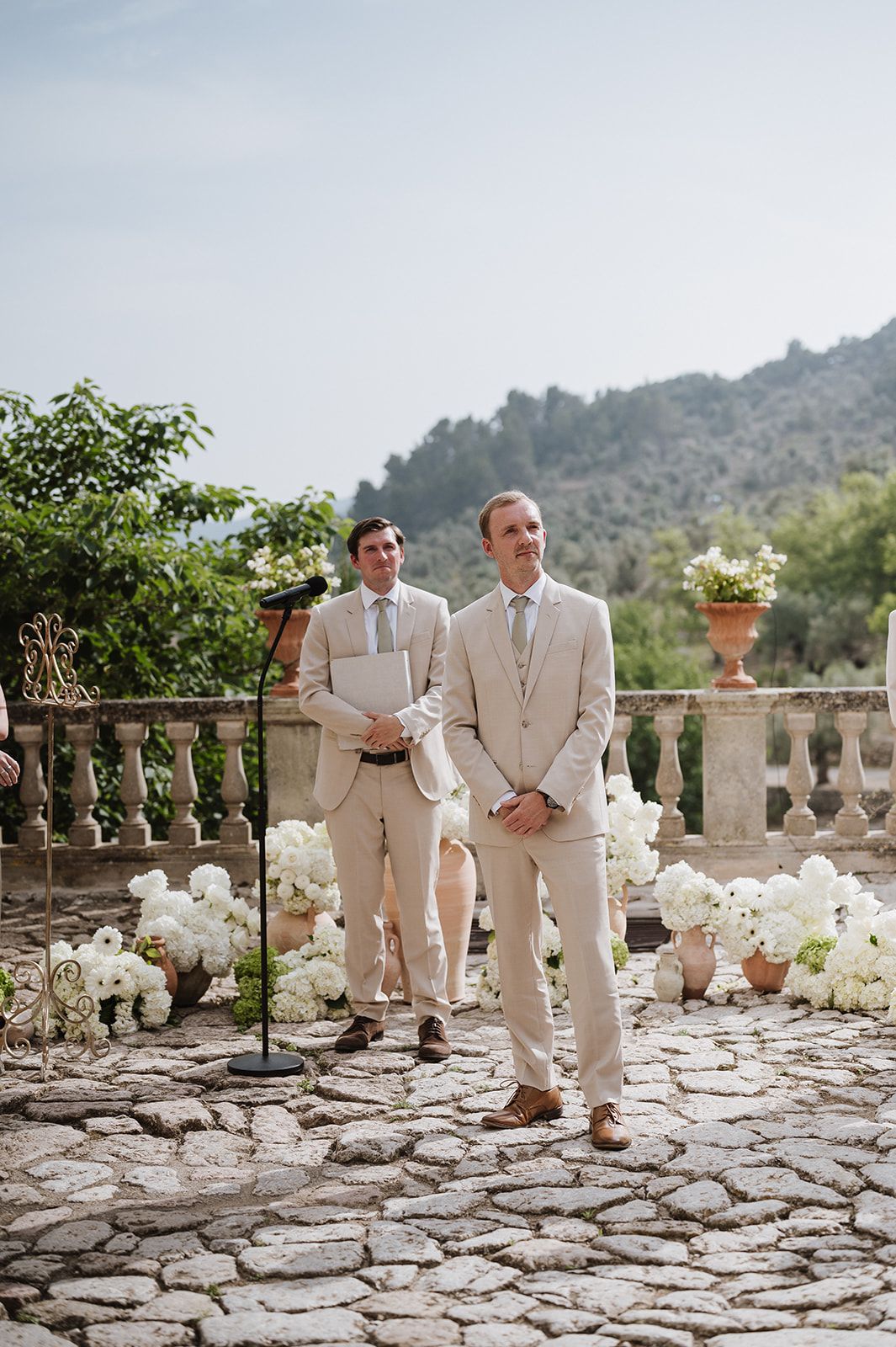 Groom in beige suit stands at outdoor wedding altar with officiant, cobblestone terrace, white floral arrangements, and mountain backdrop.