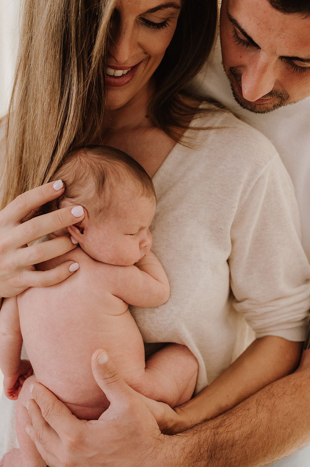 Happy parents in neutral tones smiling while carefully holding their sleeping newborn baby together.