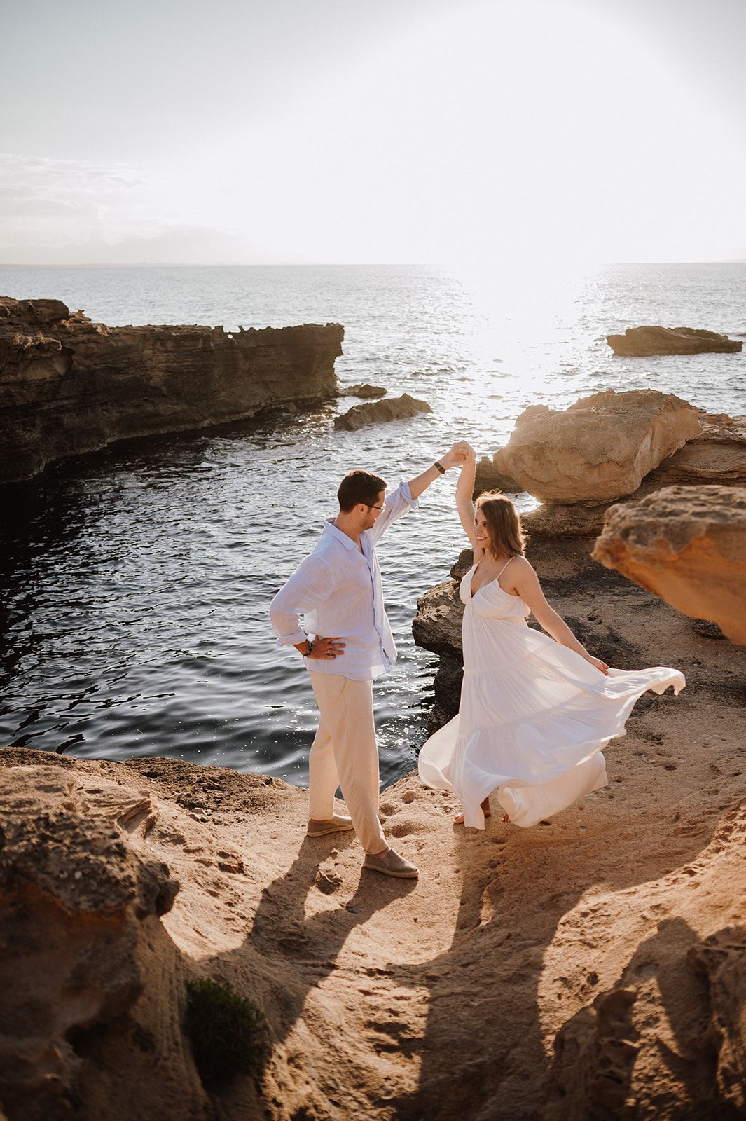 Couple dancing in white outfits on rocky coastal cliffs at golden sunset with ocean backdrop.