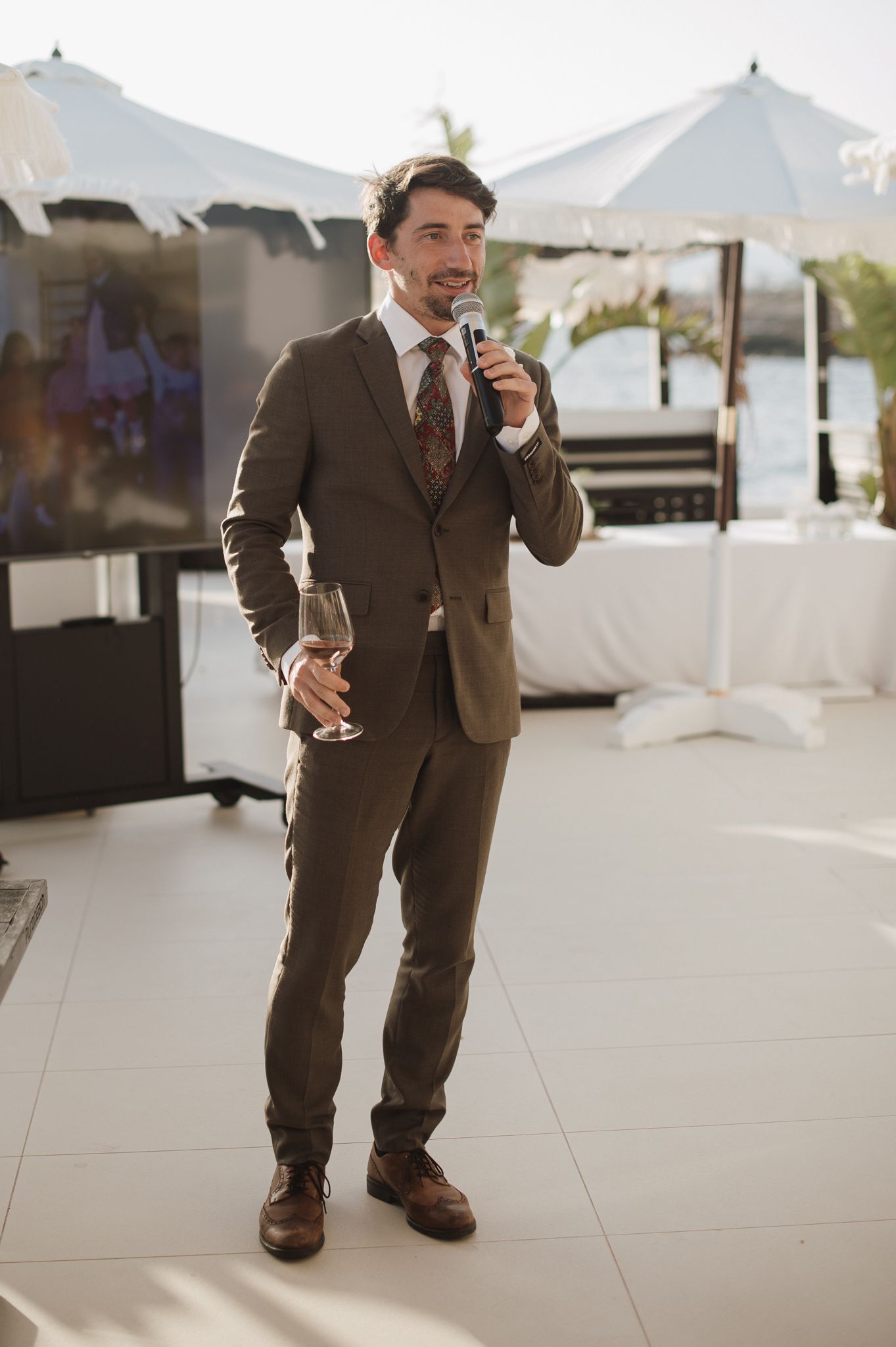 Man in brown suit gives wedding speech holding microphone and glass of wine outdoors.