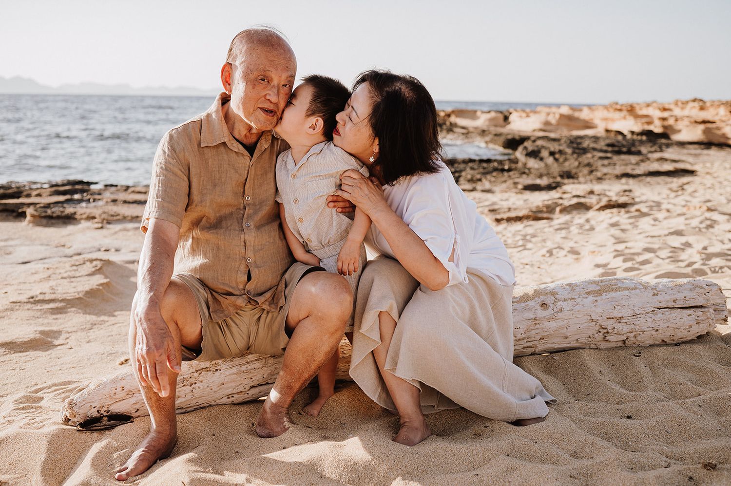 Grandparents kissing their grandson on the cheeks while sitting on a sunny sandy beach.