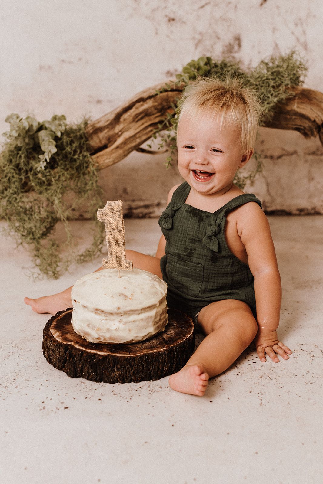 Laughing baby in green romper sits beside a first birthday smash cake on a wood slice.