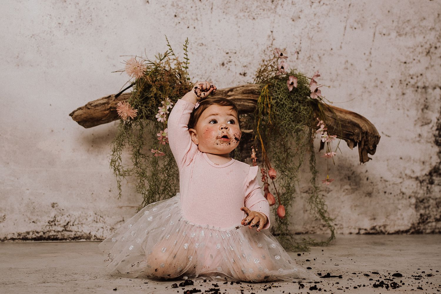 Adorable baby girl in pink tutu and bodysuit sitting in front of rustic floral backdrop for first birthday photoshoot.