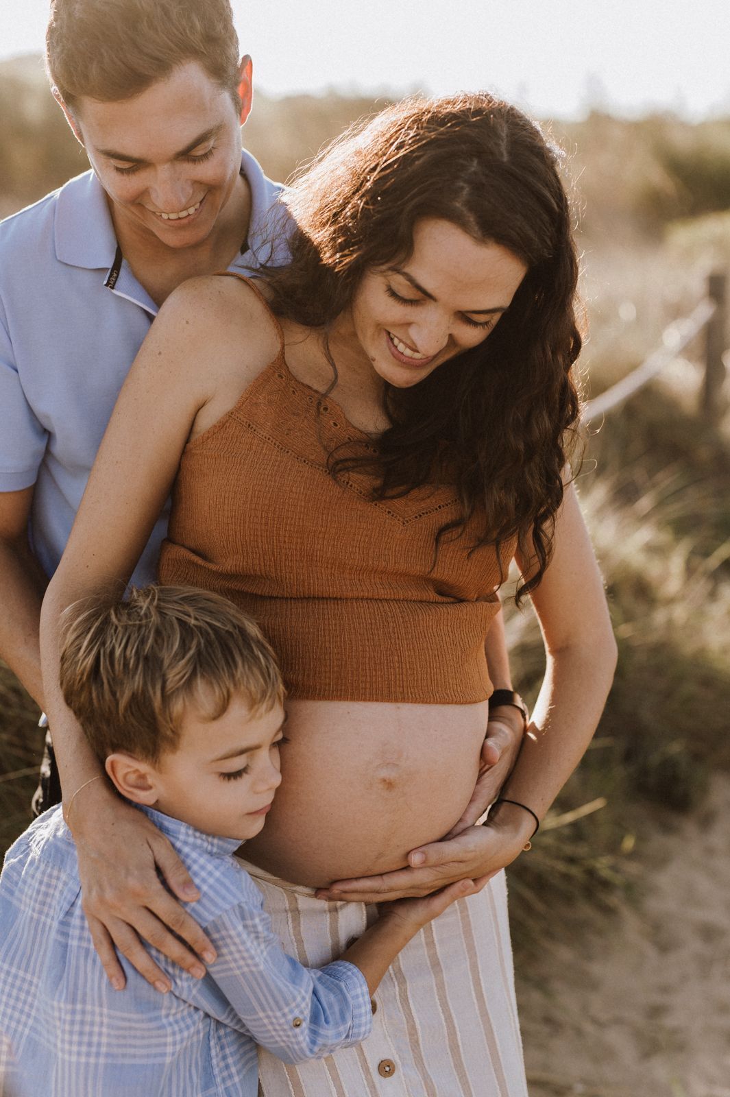 Pregnant woman smiling outdoors with husband and young son hugging her baby bump during a sunny maternity photo session.