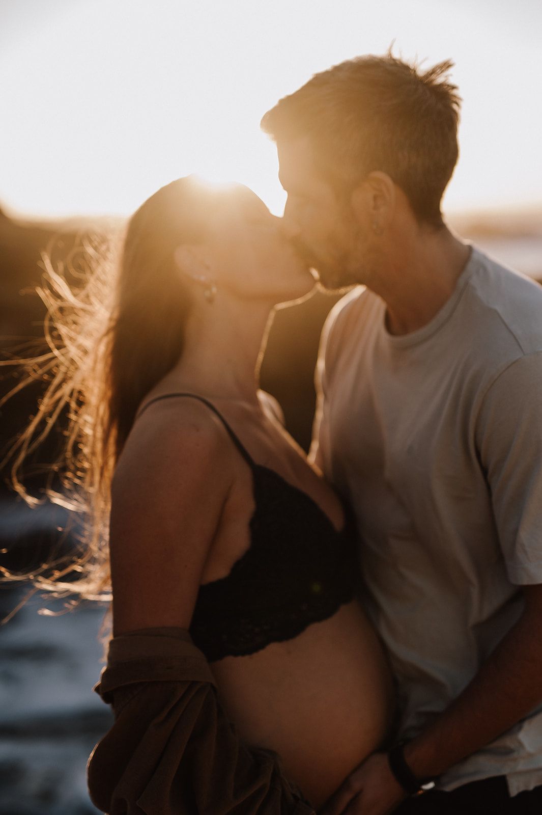 Couple sharing a romantic kiss at golden hour sunset on a beach, woman showing pregnant belly in black bikini top.