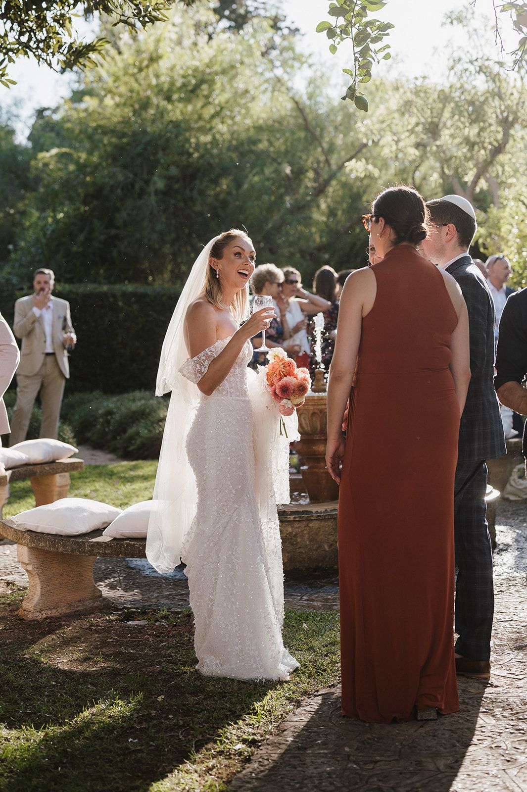 Smiling bride in white sequin gown and veil holds bouquet while chatting with guests at outdoor wedding reception.
