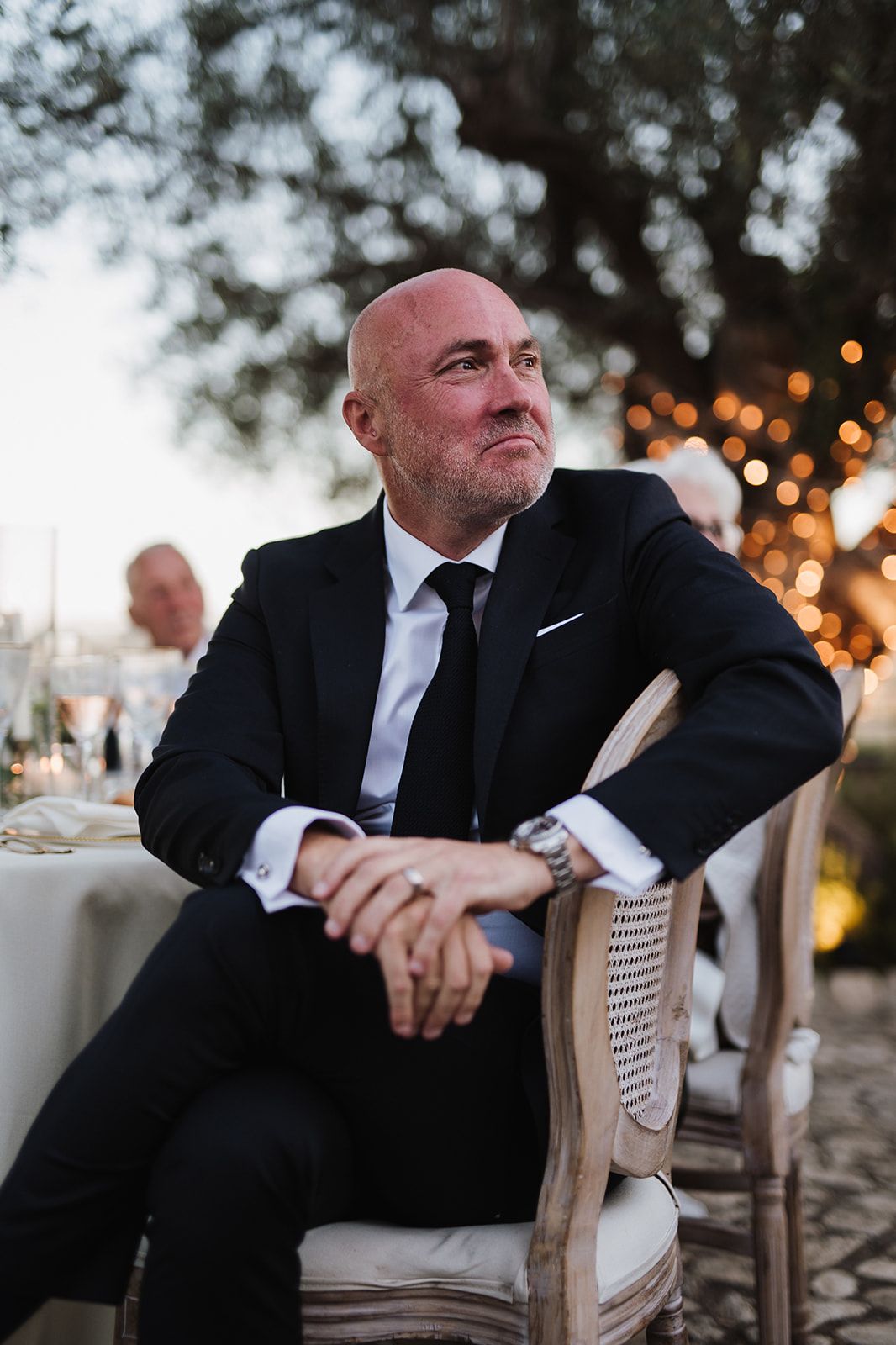 Bald man in black suit sits thoughtfully at an outdoor wedding reception under twinkling lights.