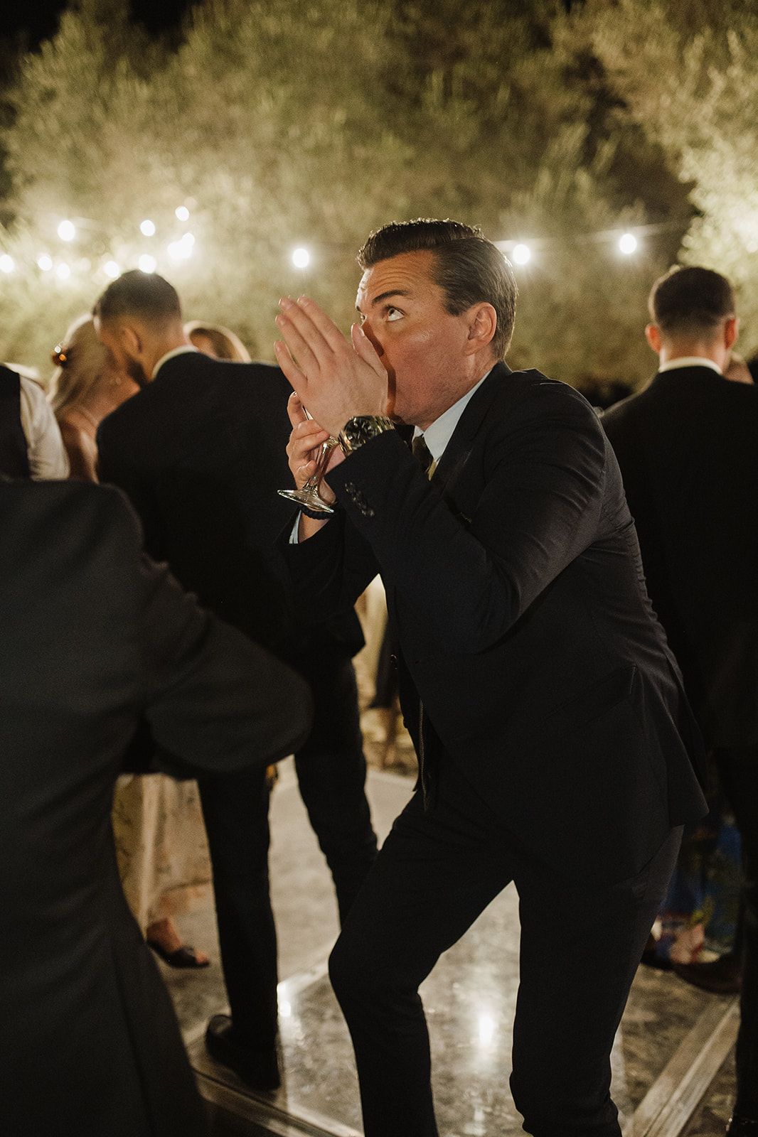 Man in black suit dancing enthusiastically at outdoor wedding reception under string lights at night.