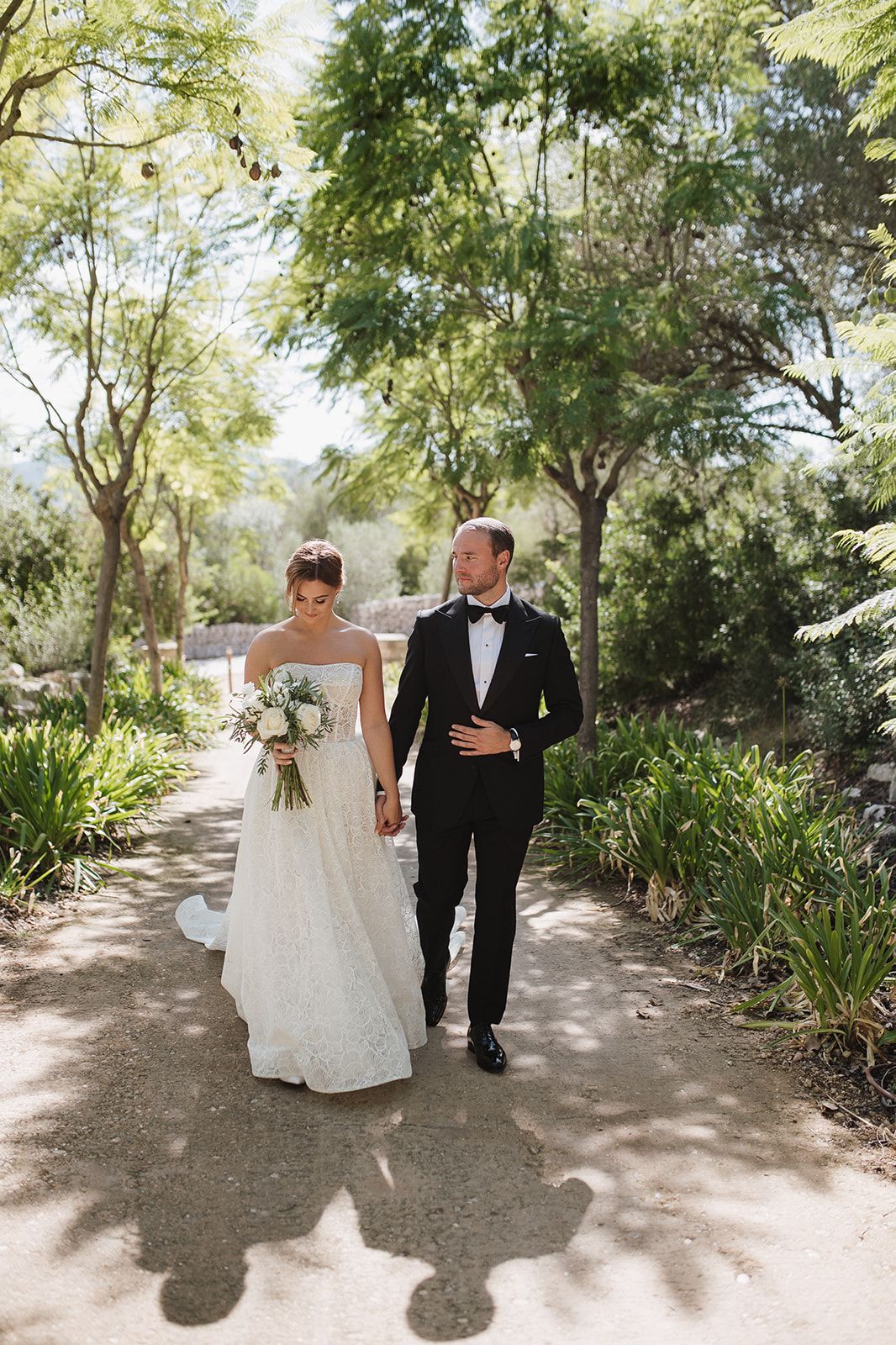 Bride and groom walk hand-in-hand along a sunlit garden path on their wedding day.