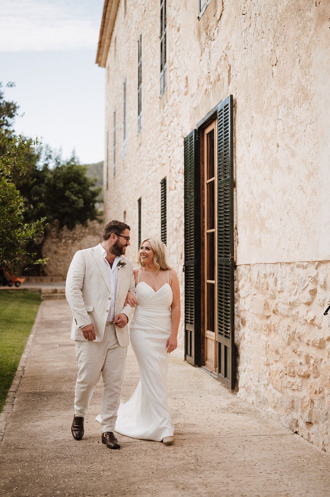 Bride and groom walk together alongside a rustic stone building, smiling at each other on their wedding day.