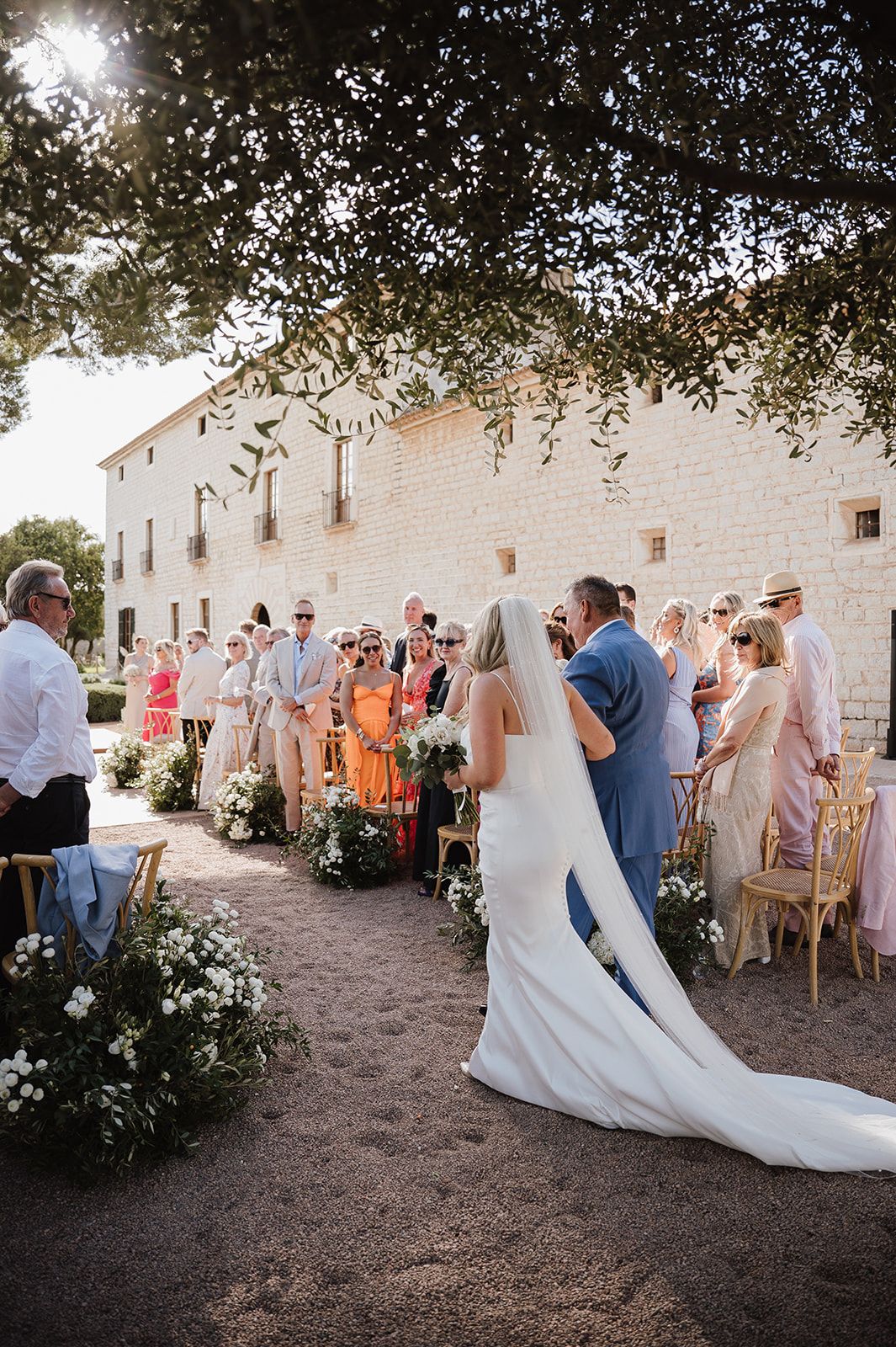 Bride walking down outdoor aisle in white gown at sunny rustic European venue.